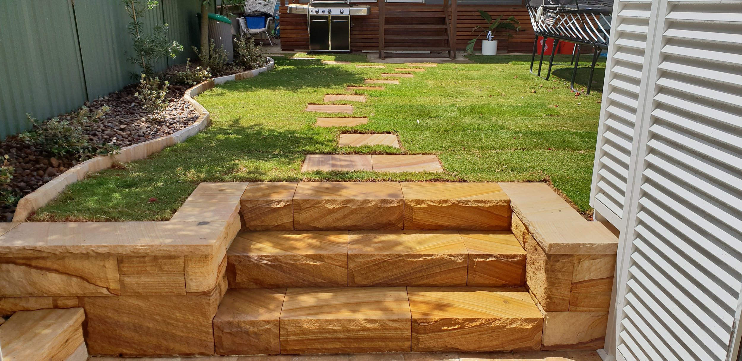 Backyard with stone steps leading down to a grassy yard, bordered by a curved flower bed with small plants and rocks, a green fence on the left, a white lattice structure on the right, and a garden area with a grill, chairs, and potted plants at the 