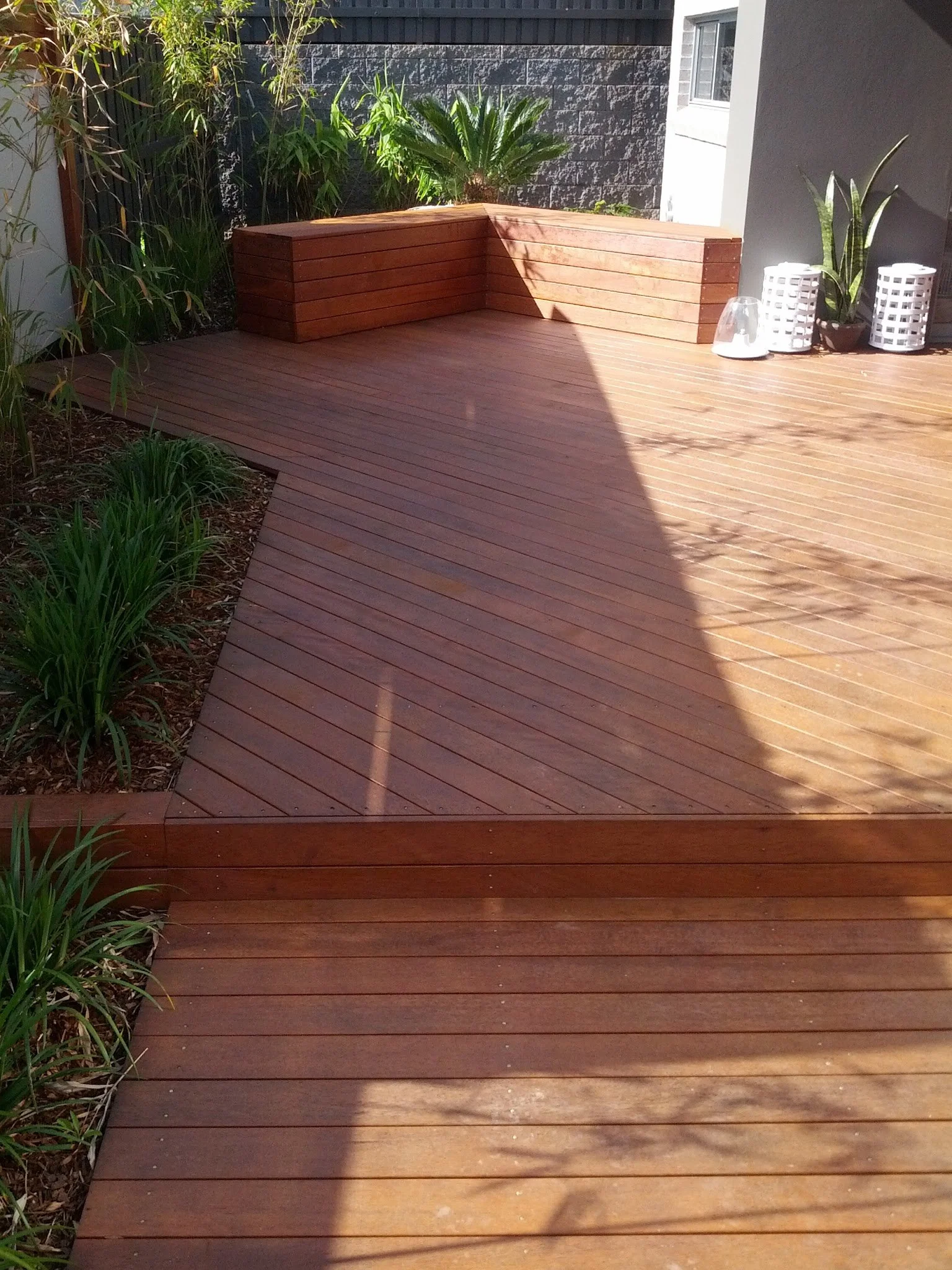 A wooden outdoor deck with built-in bench seating against a gray brick wall, plants on the left and right sides, and white decorative lanterns on the right.