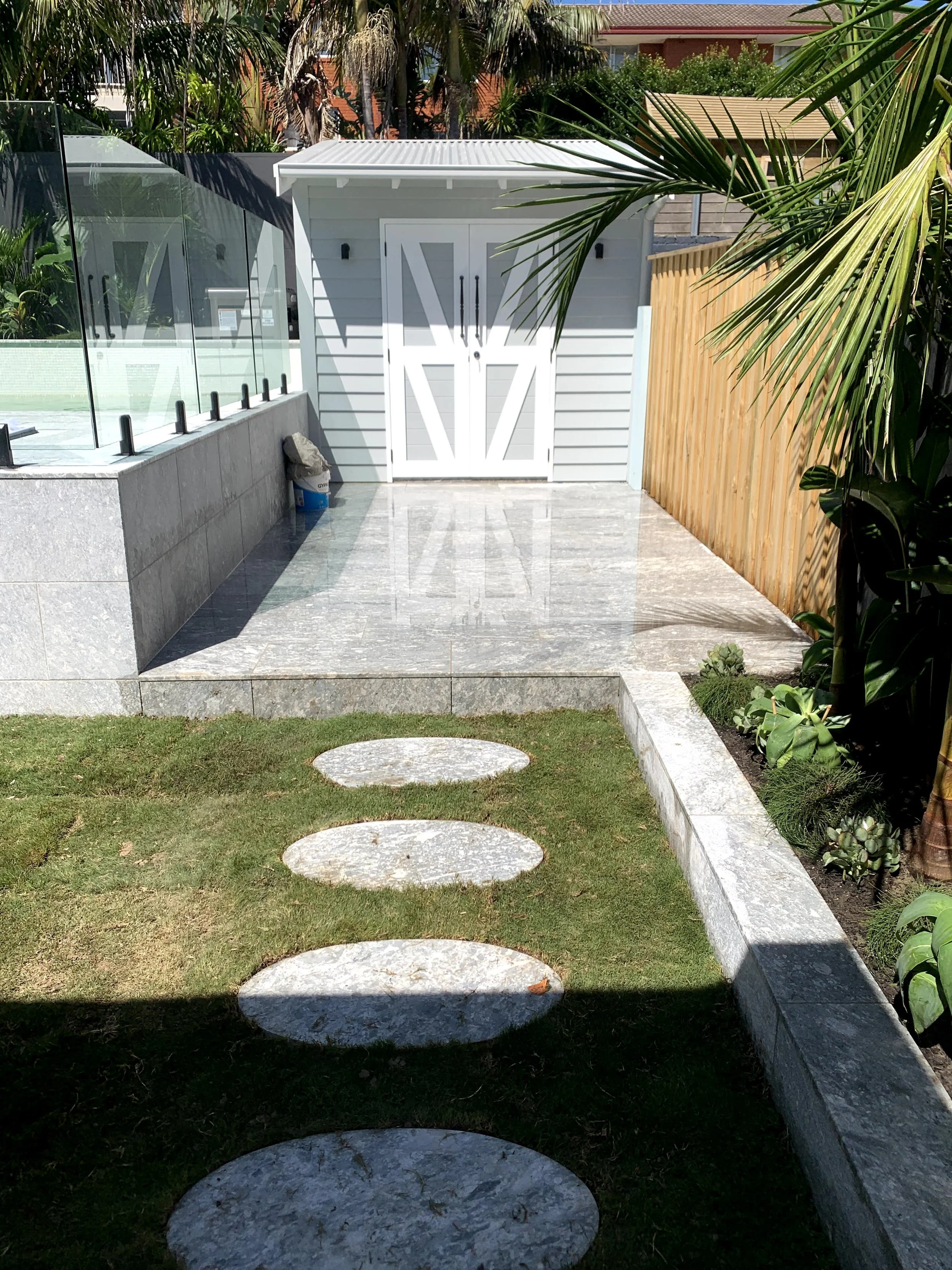 A backyard with a small white shed, a paved walkway with round stepping stones, green grass, and a garden with various plants and trees. There is a wooden fence on the right side and a glass barrier on the left, with a concrete ledge and some gardeni