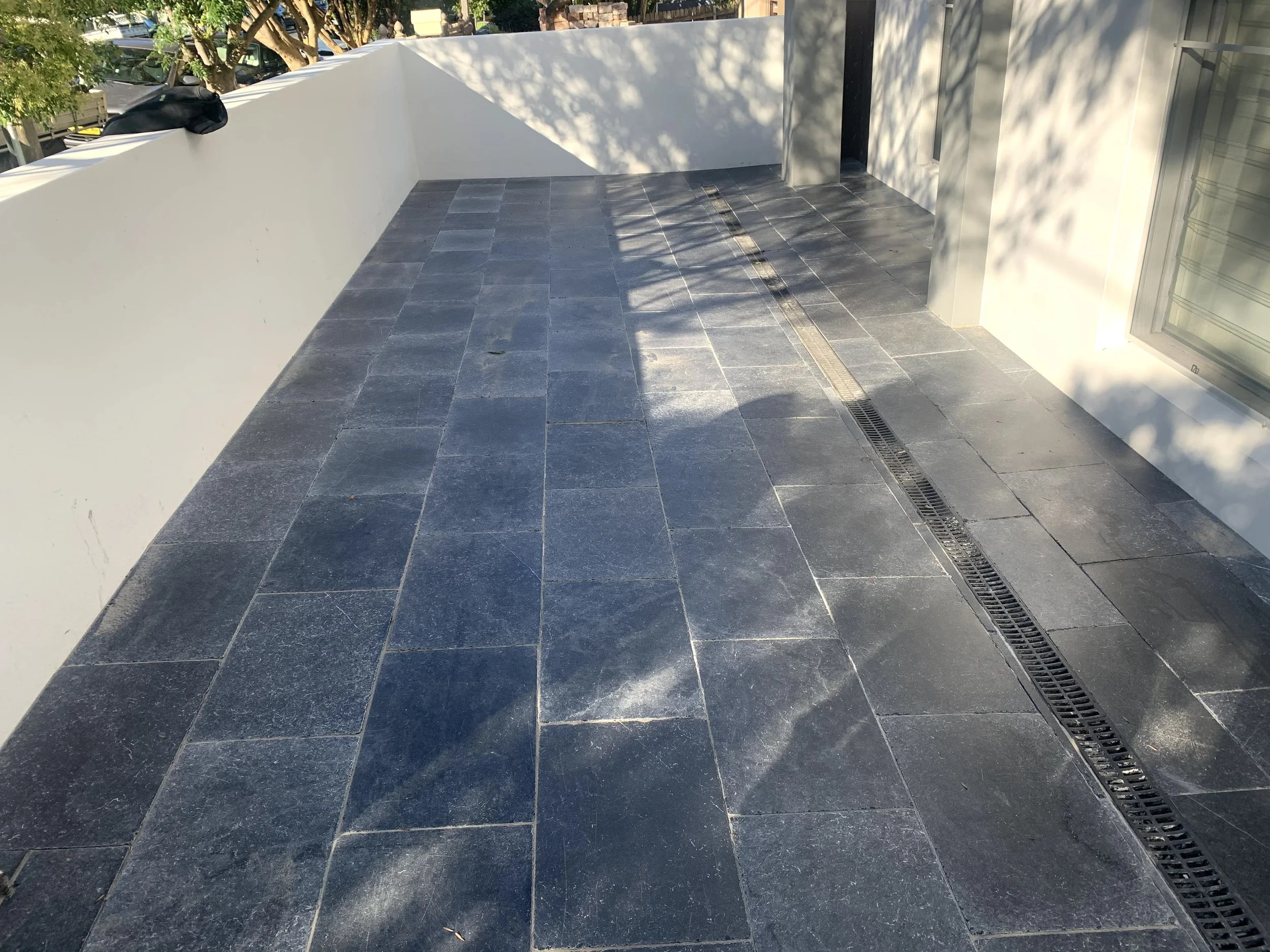 A modern outdoor balcony with dark gray stone tiles, a white wall and railing, and a narrow drain grate along the edge. Shadows of nearby trees cast patterns on the tiles.