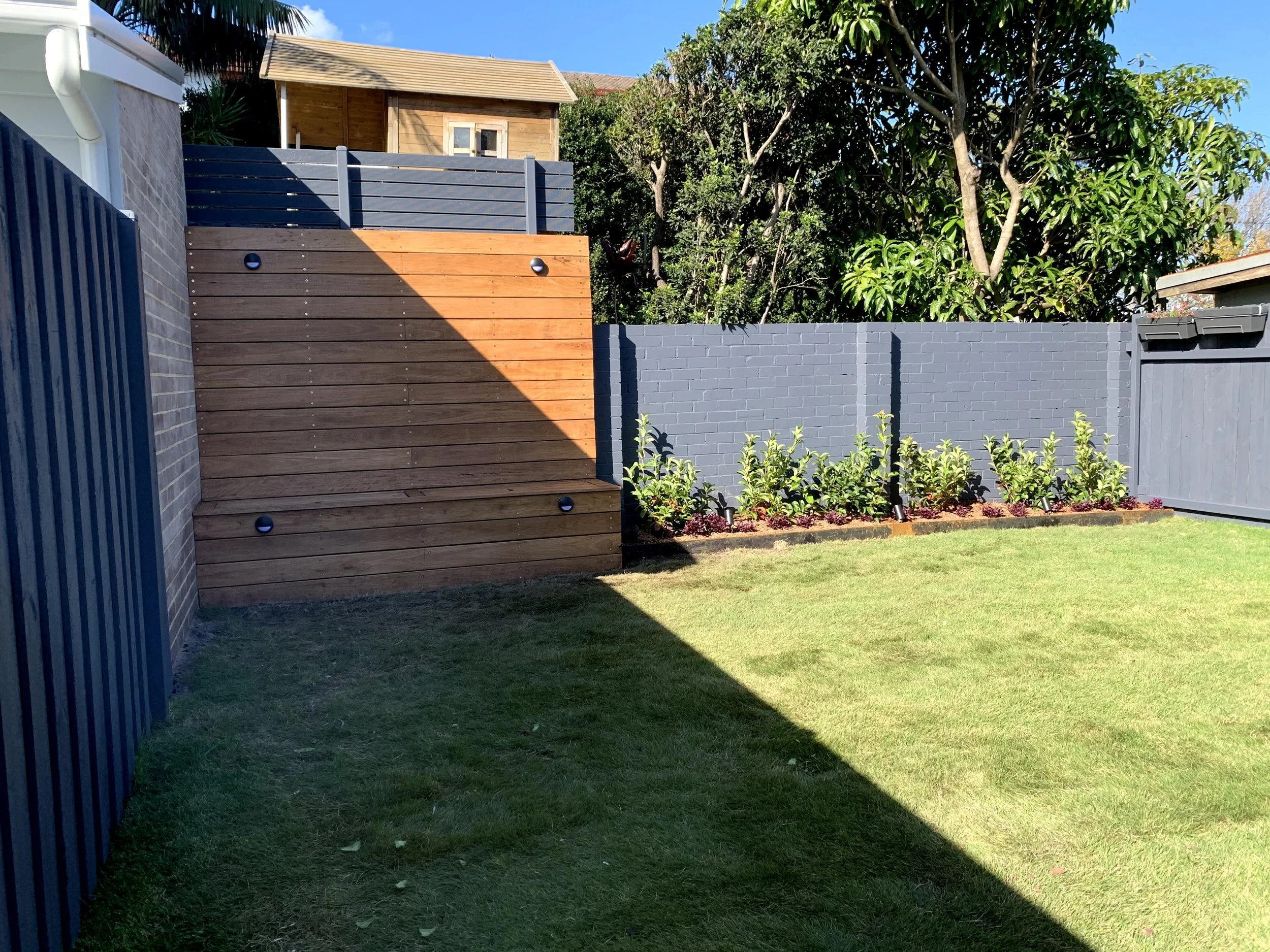 A backyard scene showing a grassy lawn, a tall dark gray brick wall, a wooden fence with two mounted lights, a small wooden shed on a raised platform, and lush leafy trees against a blue sky.