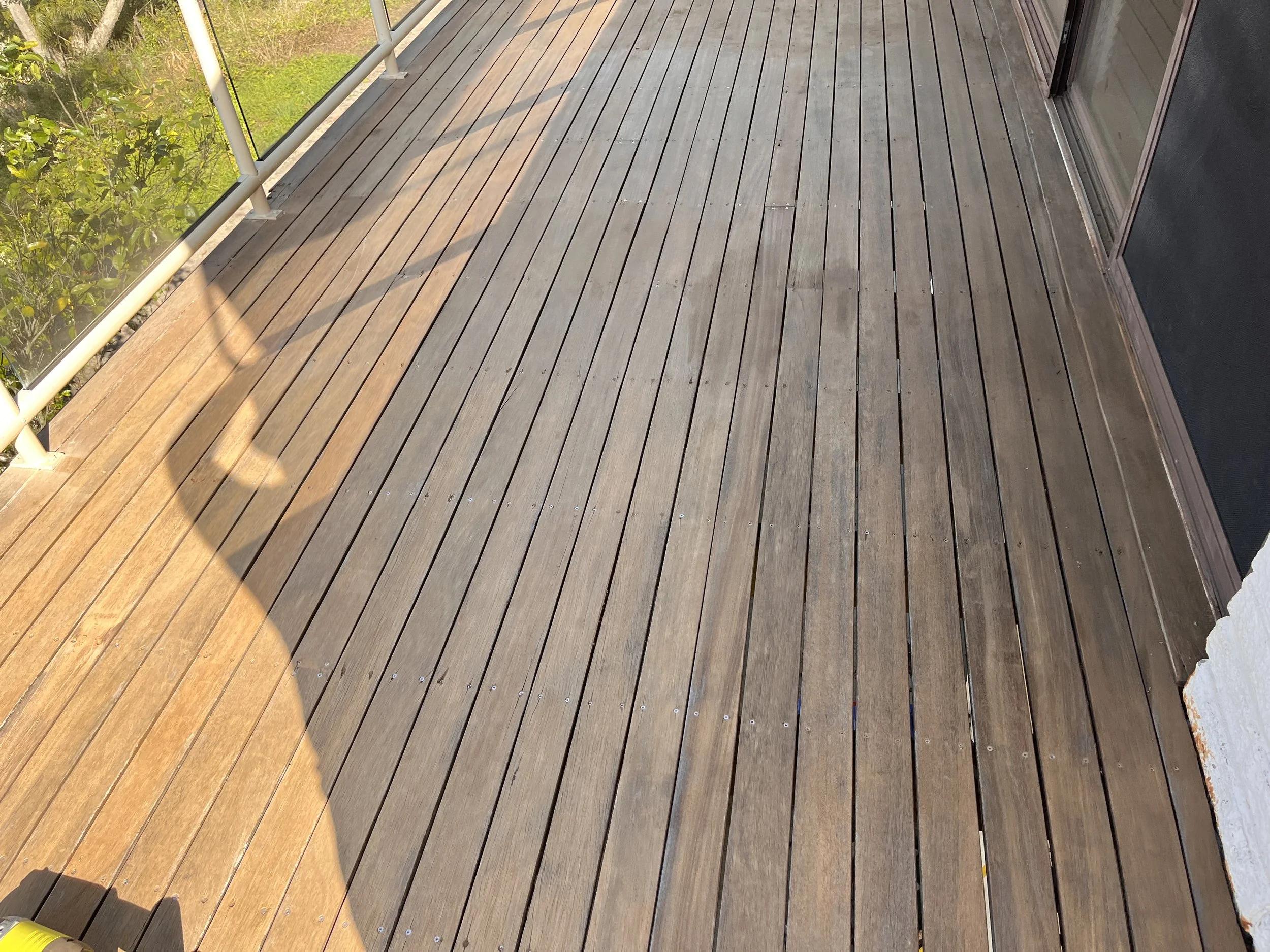 An empty outdoor wooden deck with a glass railing on the left side and a sliding door on the right side.