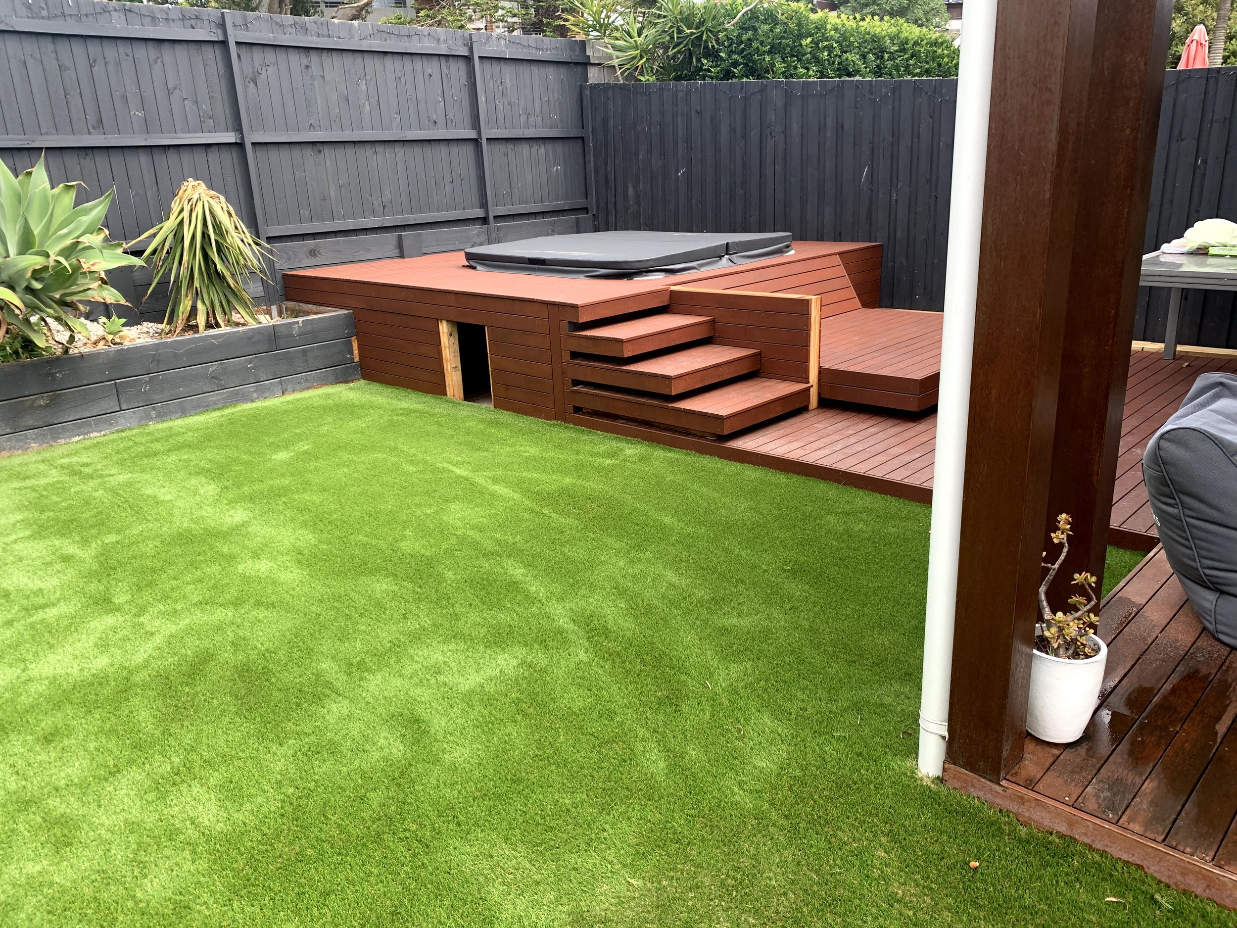 A backyard with a green grass lawn, a raised wooden deck with stairs, and a hot tub cover on the deck. There are plants and shrubs along a black wooden fence that surrounds the yard. Part of a table with some cloth and other items is visible on the r