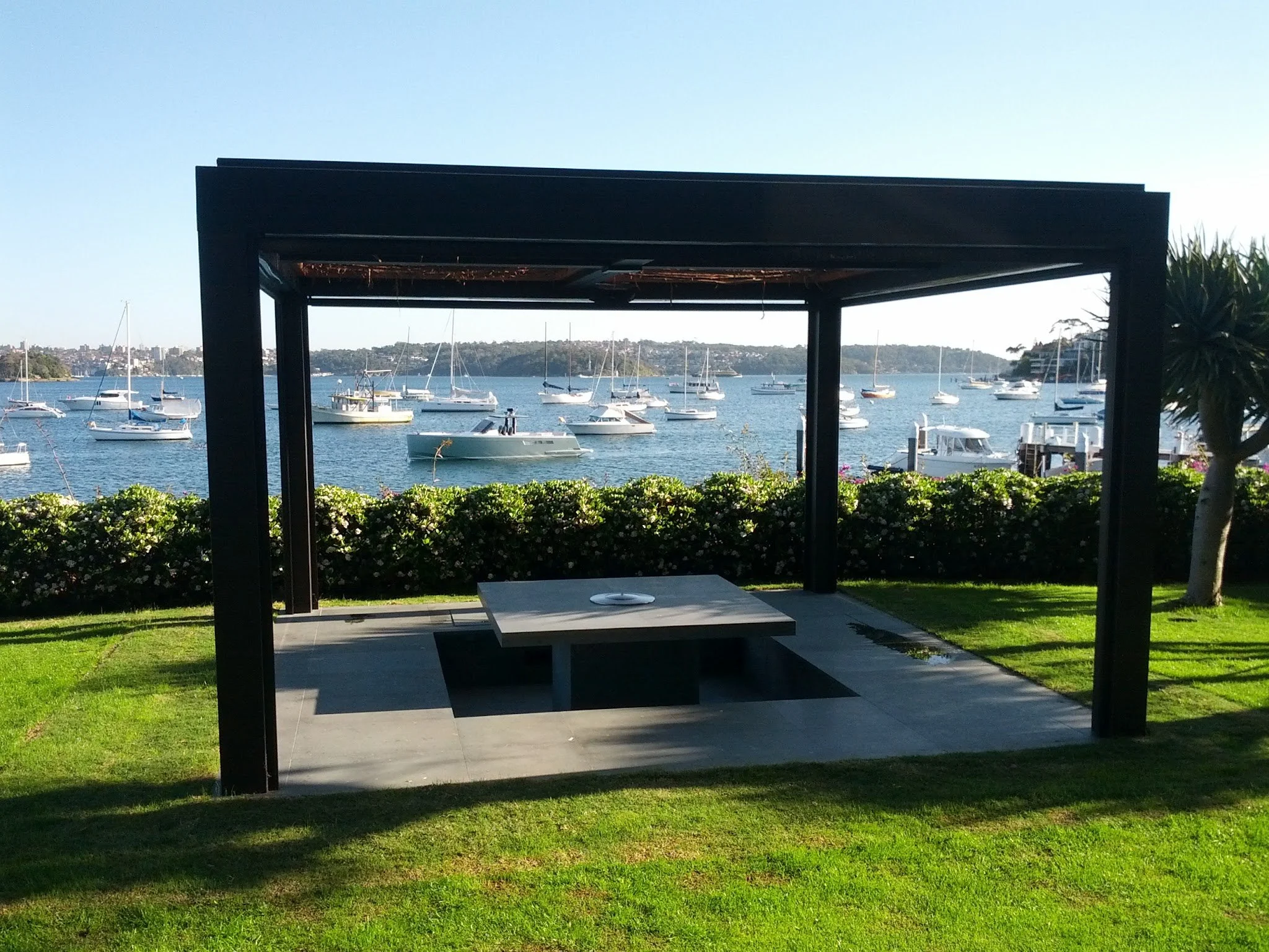 A lakeside view with multiple sailboats and yachts anchored on the water, seen through a dark-colored pergola structure on a grassy area.