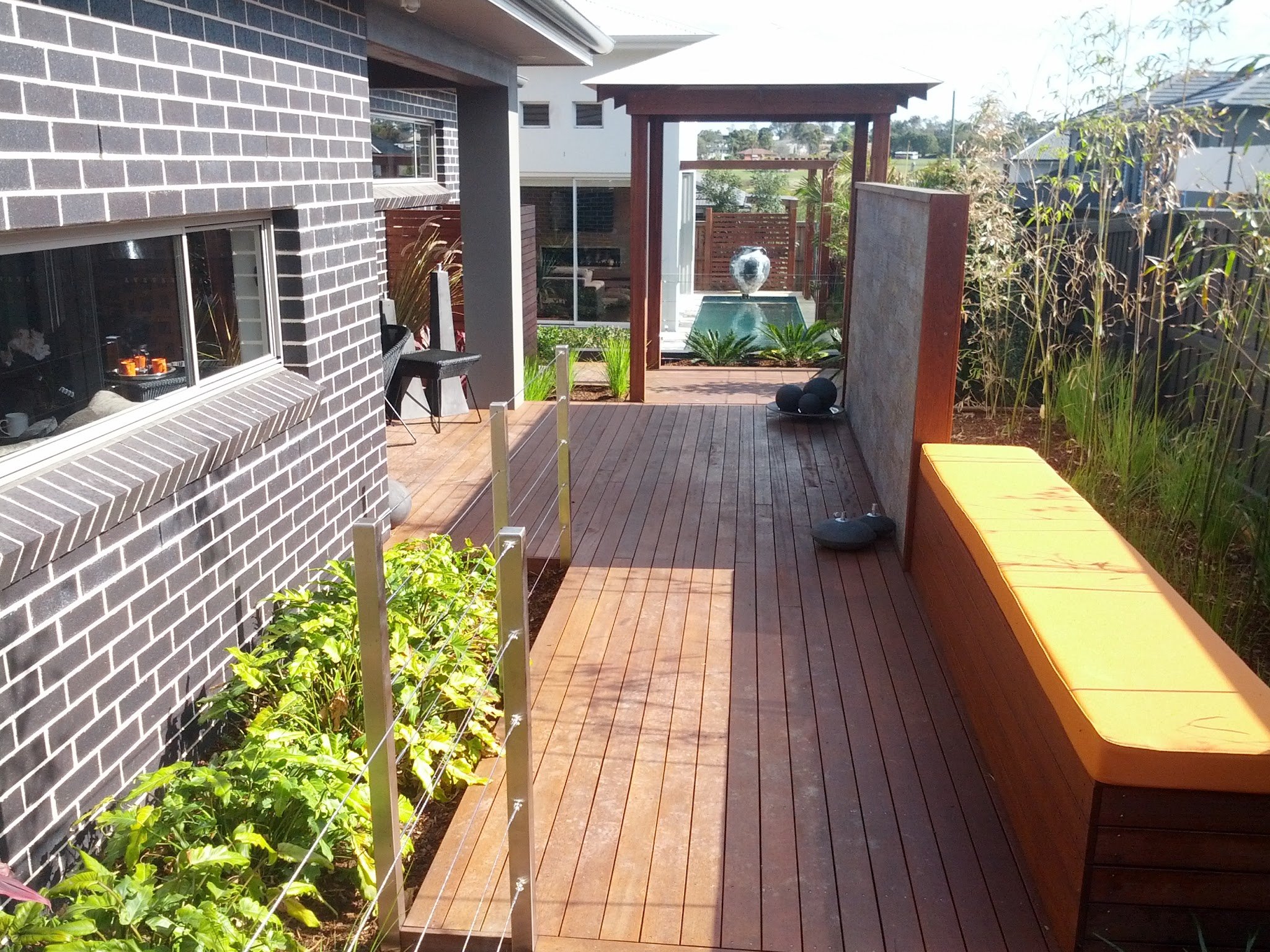 A backyard patio with a wooden deck, a yellow bench, a brick house with a window, and a water fountain with a sculpture, surrounded by plants and trees, under a bright sky.
