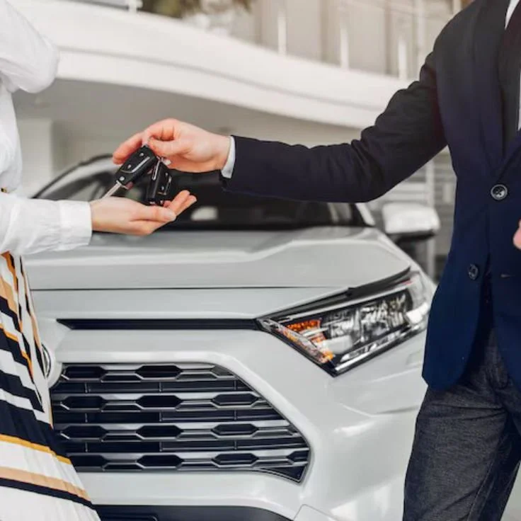 A man in a suit hands over car keys to a woman in front of a white car.