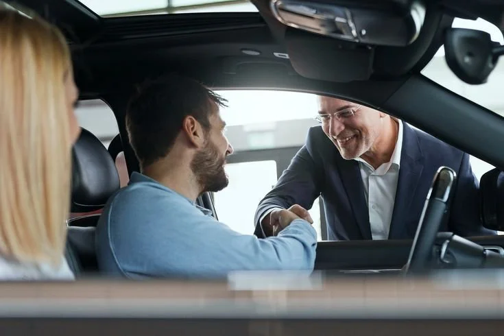 A man in a suit shaking hands with a customer inside a car dealership.