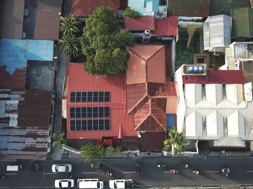 An aerial view of a street with several buildings, the central one has a red roof with solar panels and surrounding trees.
