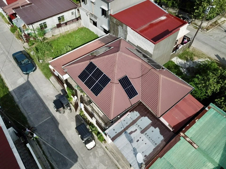 An aerial view of a residential neighborhood showing houses with solar panels on their roofs, parked cars on the street, and trees and greenery around.