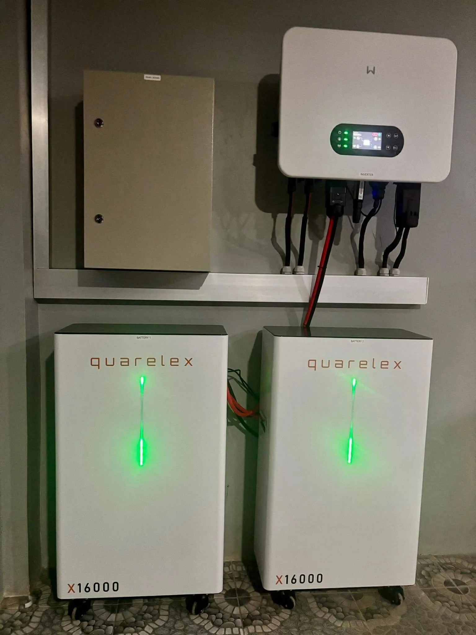 Two Quarelex batteries with a white inverter mounted on the wall above them. Both batteries are labeled with a price of 16,000. The batteries have green lights and a vertical green line on each. The inverter has a digital display and multiple indicator lights.