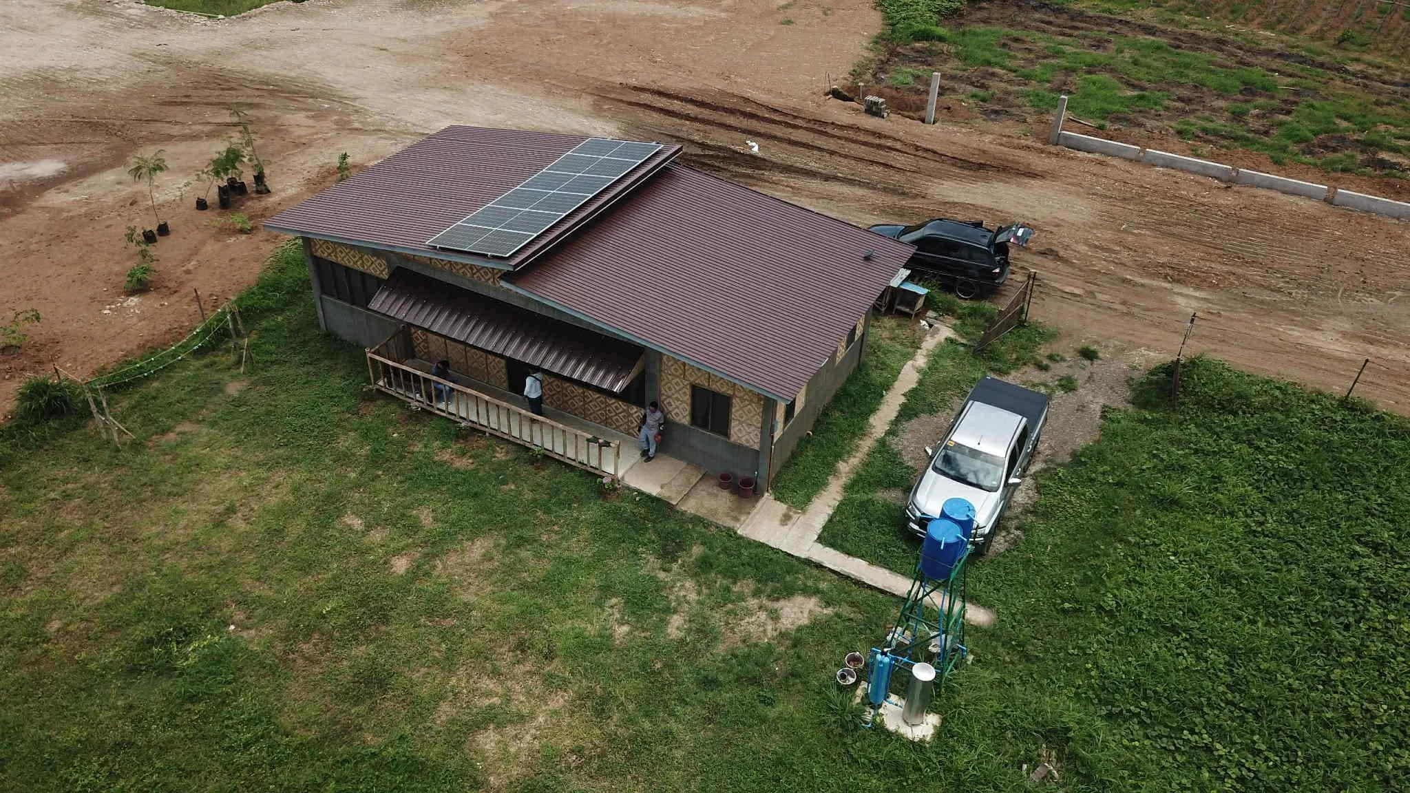 Aerial view of a house with solar panels on its roof, surrounded by a garden, parked cars, and a dirt road in a semi-rural area.