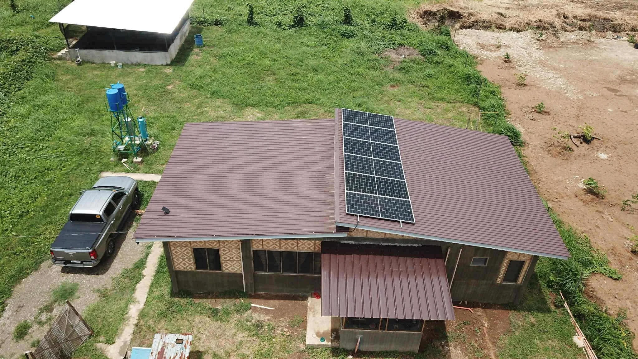A house with solar panels on the roof, a parked pickup truck, a water tower, a grassy yard, and a section of bare dirt ground.
