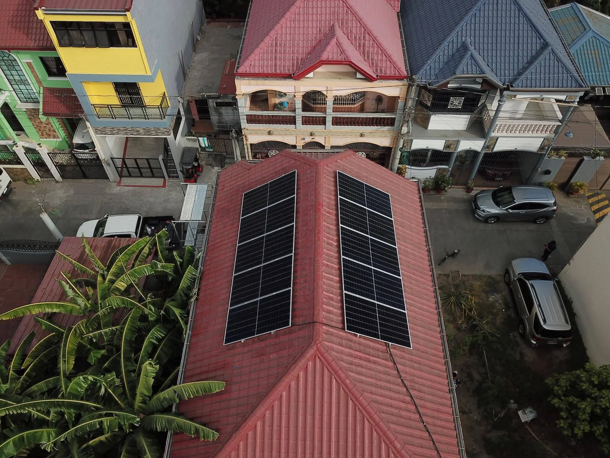 Aerial view of a row of houses with red, blue, and colored roofs, some of which have solar panels installed. Cars are parked on the street in front of the houses, and there are trees and plants visible along the street.