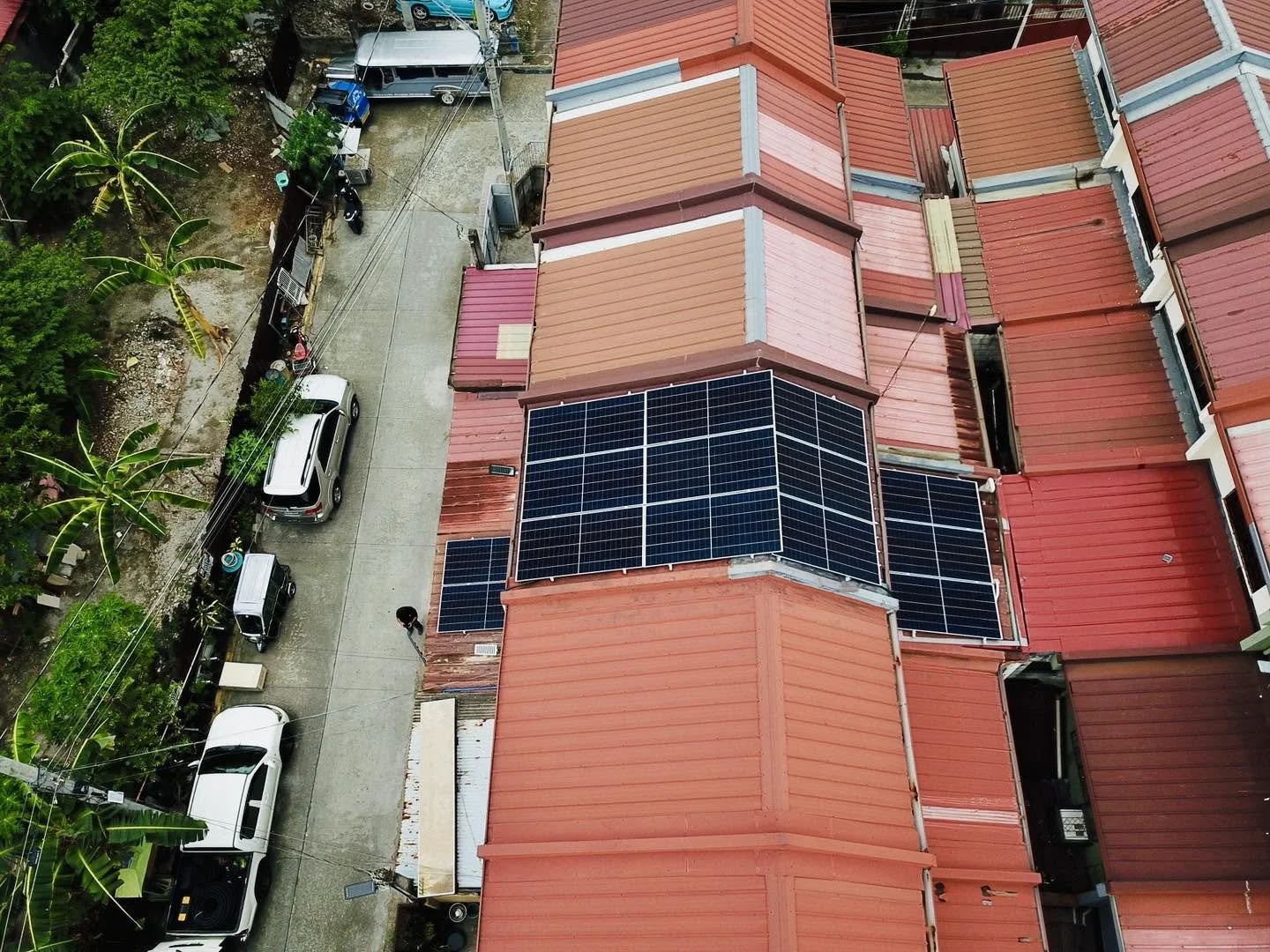 Aerial view of a neighborhood with red rooftops, featuring solar panels on one roof, a narrow street with parked cars, trees, and power lines.