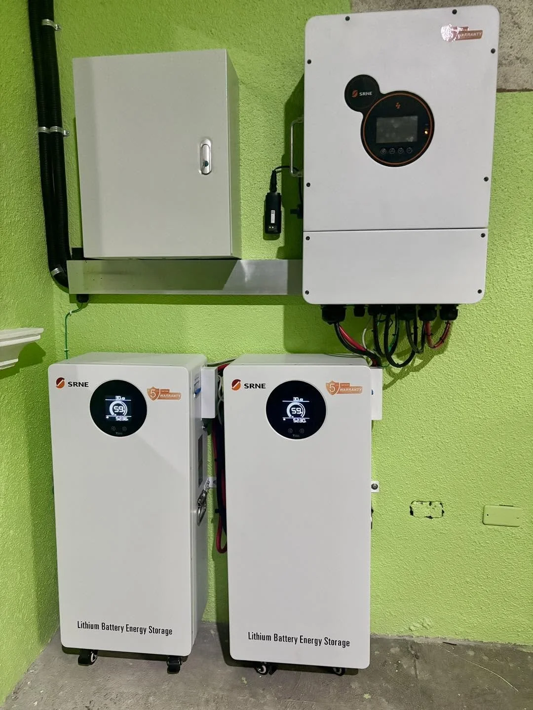 Two lithium battery energy storage units with digital displays, connected to an electrical panel on a bright green wall.