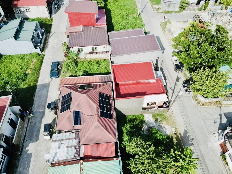 An aerial view of a neighborhood with several houses, some with solar panels on the roofs, surrounded by trees, grass, and streets with parked cars.