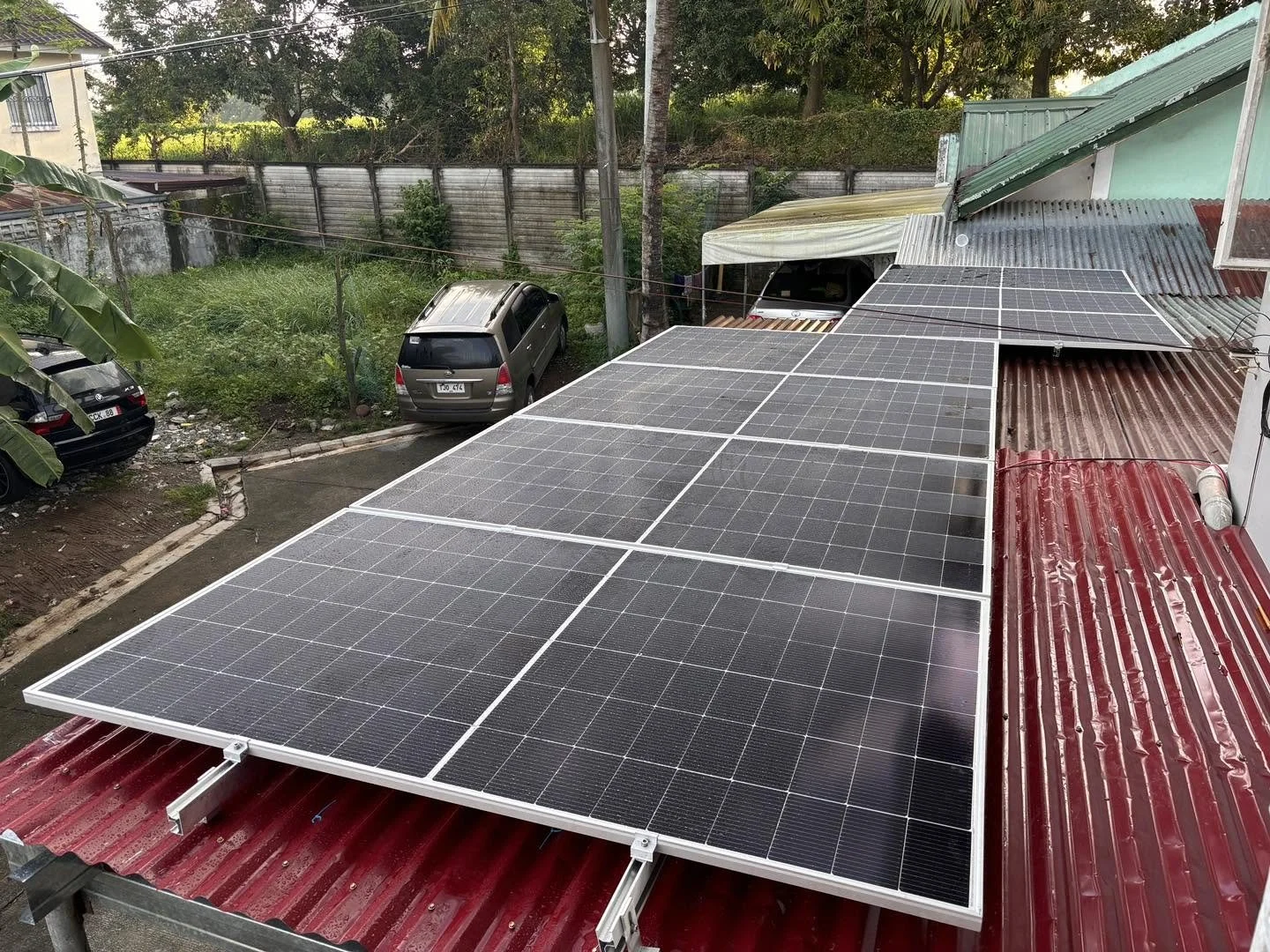 A rooftop with multiple solar panels installed, red roofing material, and a residential area with cars parked nearby and greenery in the background.