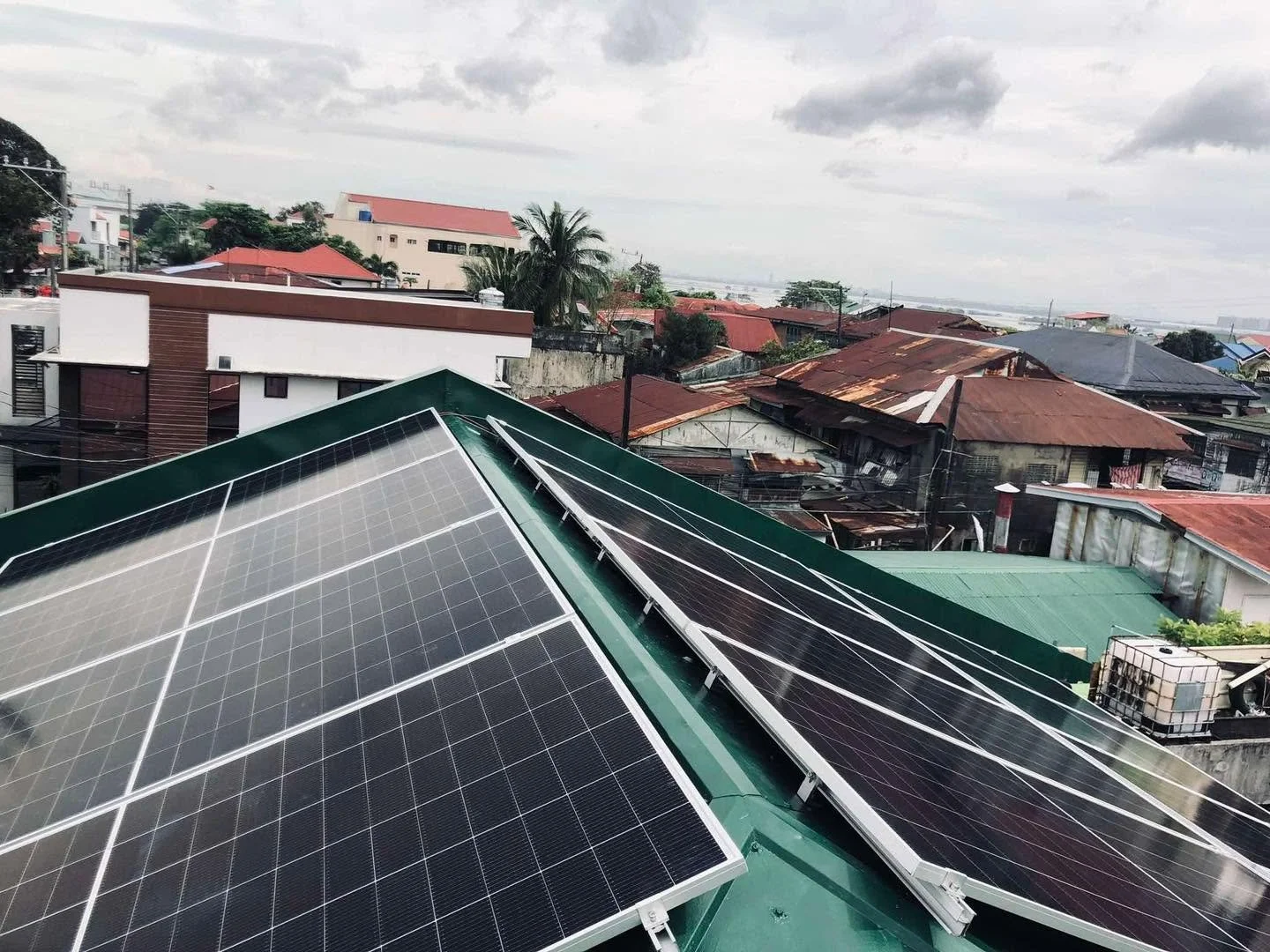 Solar panels installed on a green rooftop with a cityscape of various houses and buildings in the background, under cloudy skies.
