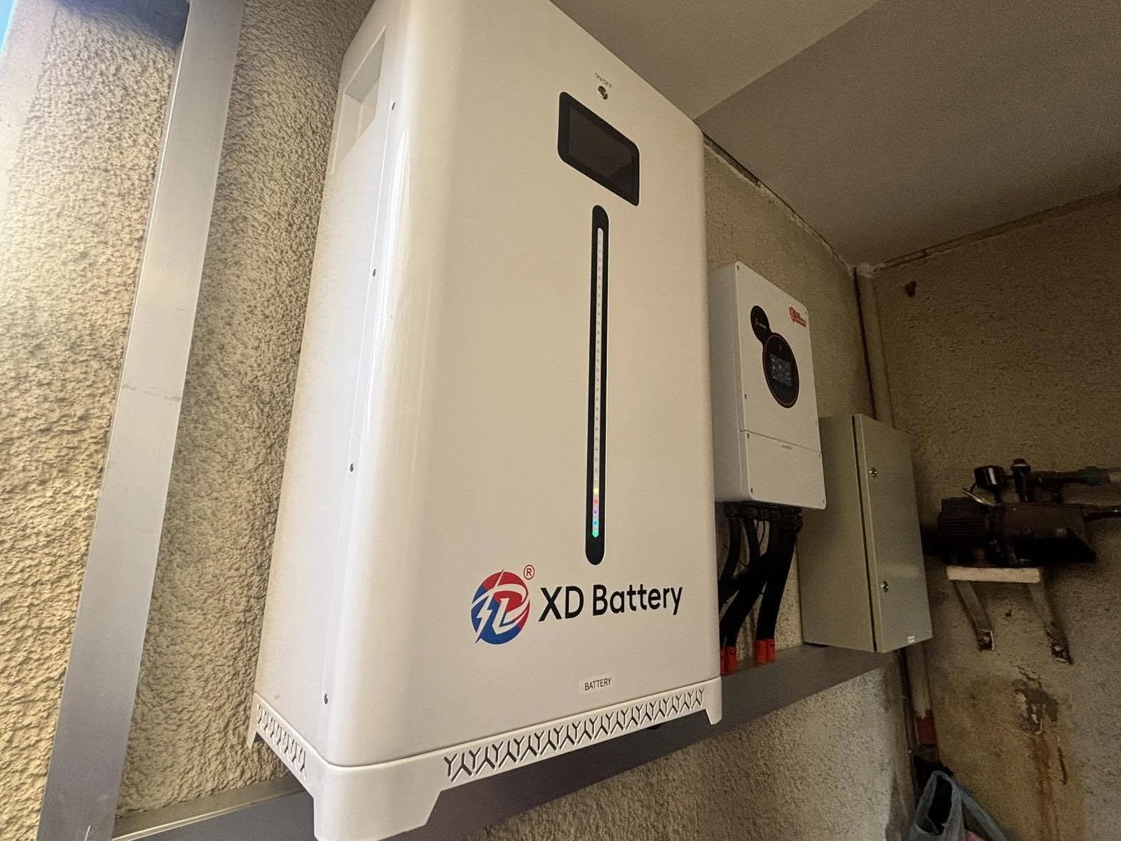 A white XD Battery storage unit with a digital display and indicator lights, mounted on a wall, adjacent to a gray electrical control box and other utility equipment.