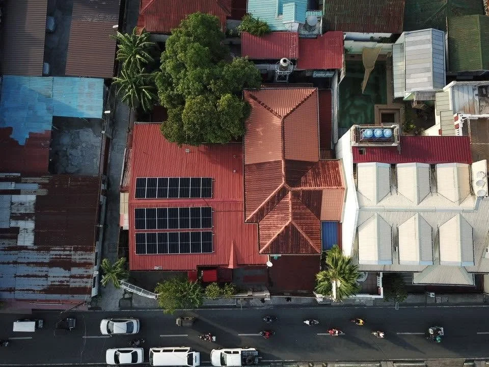 An aerial view of a neighborhood showing multiple rooftops, some with solar panels, a prominent red roof with a tree nearby, and a street with parked and moving cars.