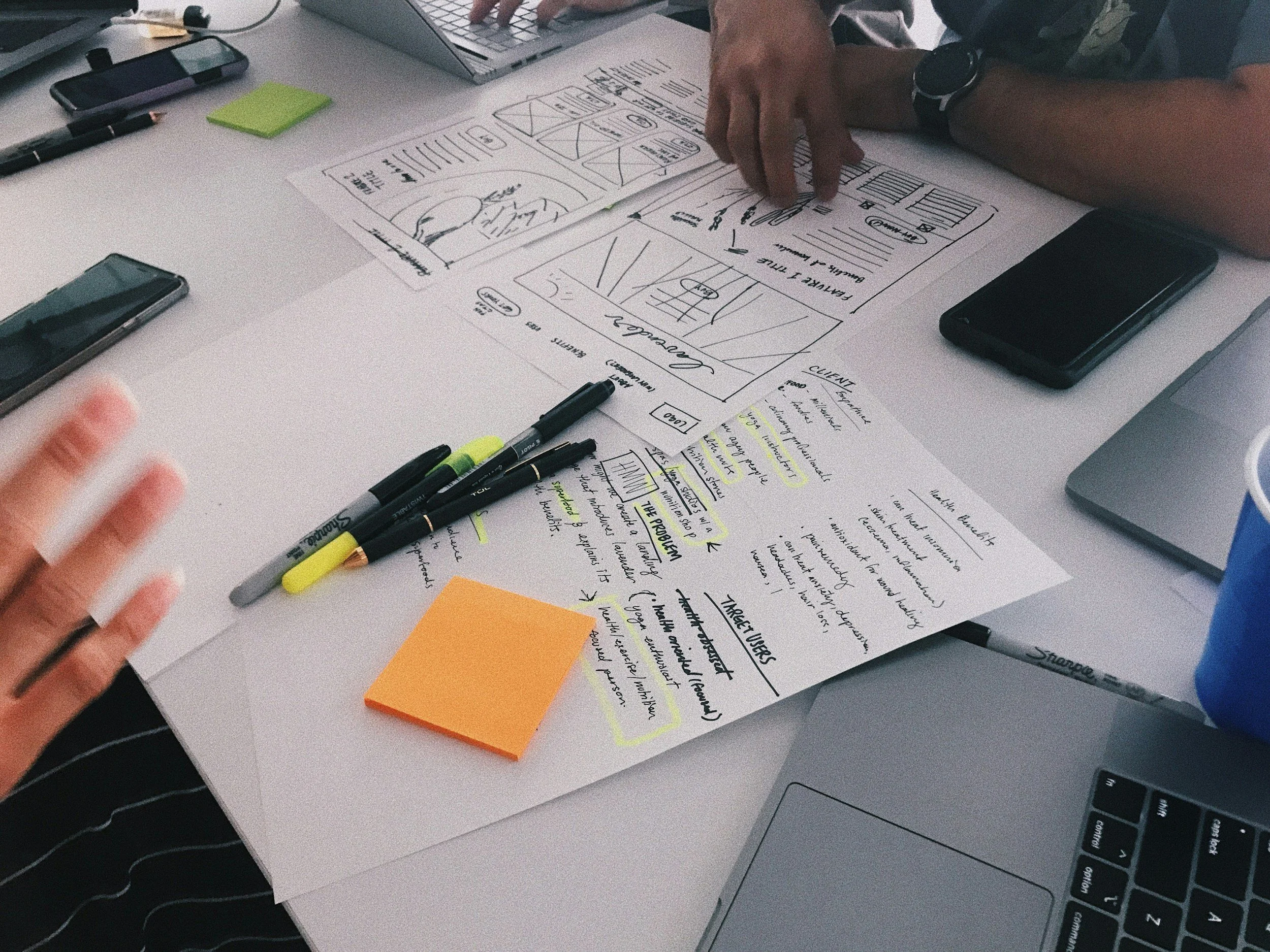 A person's arms and hands showing are working on a desk with a sketch of a mobile app or website layout, surrounded by pens, highlighters, smartphones, a laptop, and a computer keyboard.