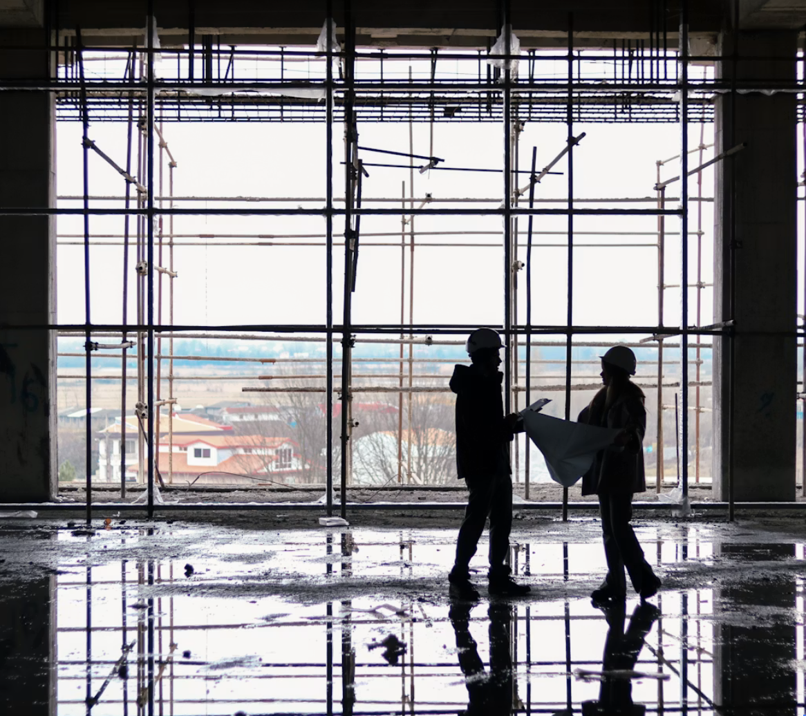 Two construction workers with helmets reviewing blueprints inside a building under construction with a large window and scaffolding in the background.