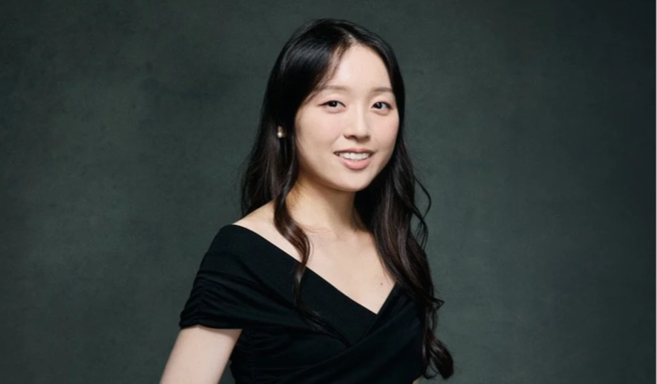 A young woman with long dark hair wearing a black top posing against a dark background.