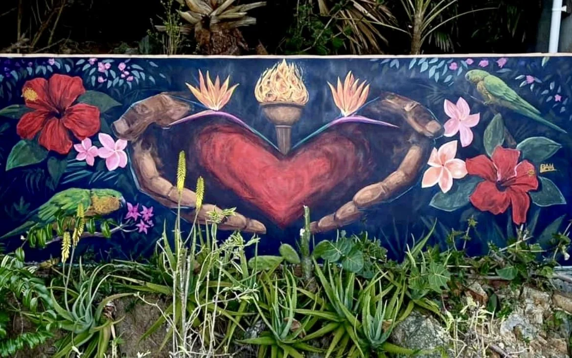 A vibrant mural depicting a large red heart surrounded by colorful flowers, tropical plants, and birds, with greenery in front.