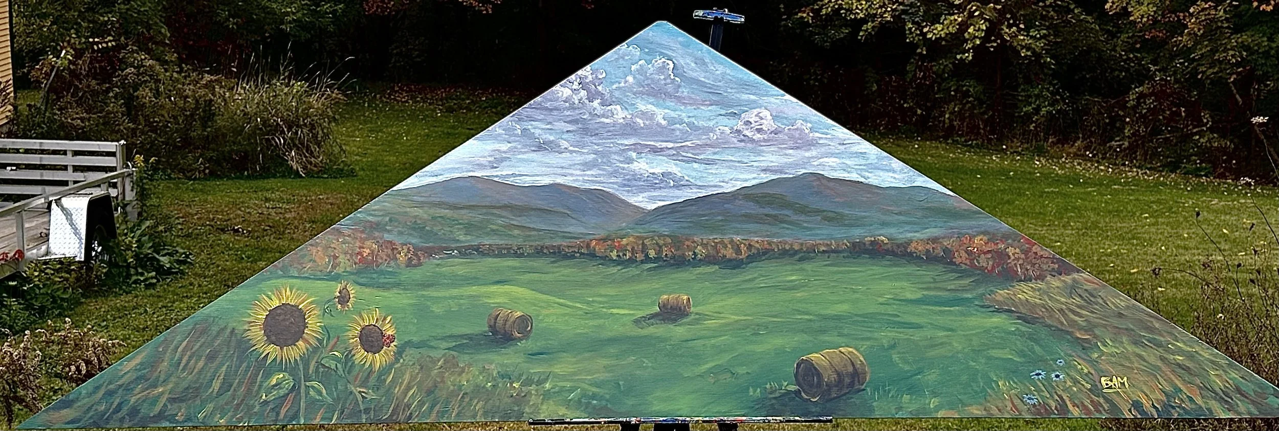A painted landscape scene on a large, flat surface outdoors, featuring mountains, clouds, a green field with sunflower and other flowers, and hay bales, with tree and shrub background.
