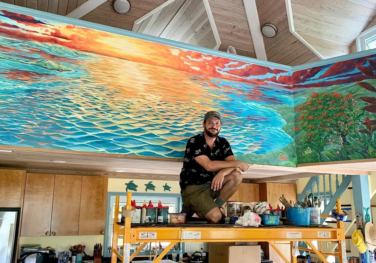 An artist squatting on a yellow scaffold in a room with wooden walls, working on a large mural of a sunset over a body of water with trees in the background.