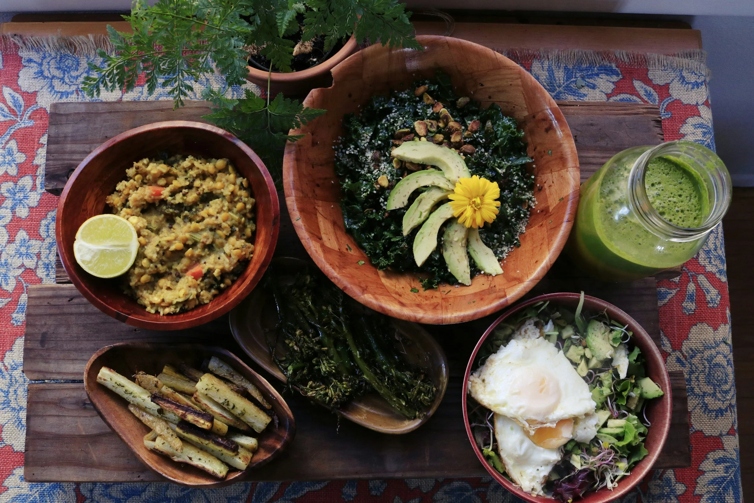 A colorful meal spread on a wooden table, featuring salads, grilled vegetables, and a green smoothie in a jar.