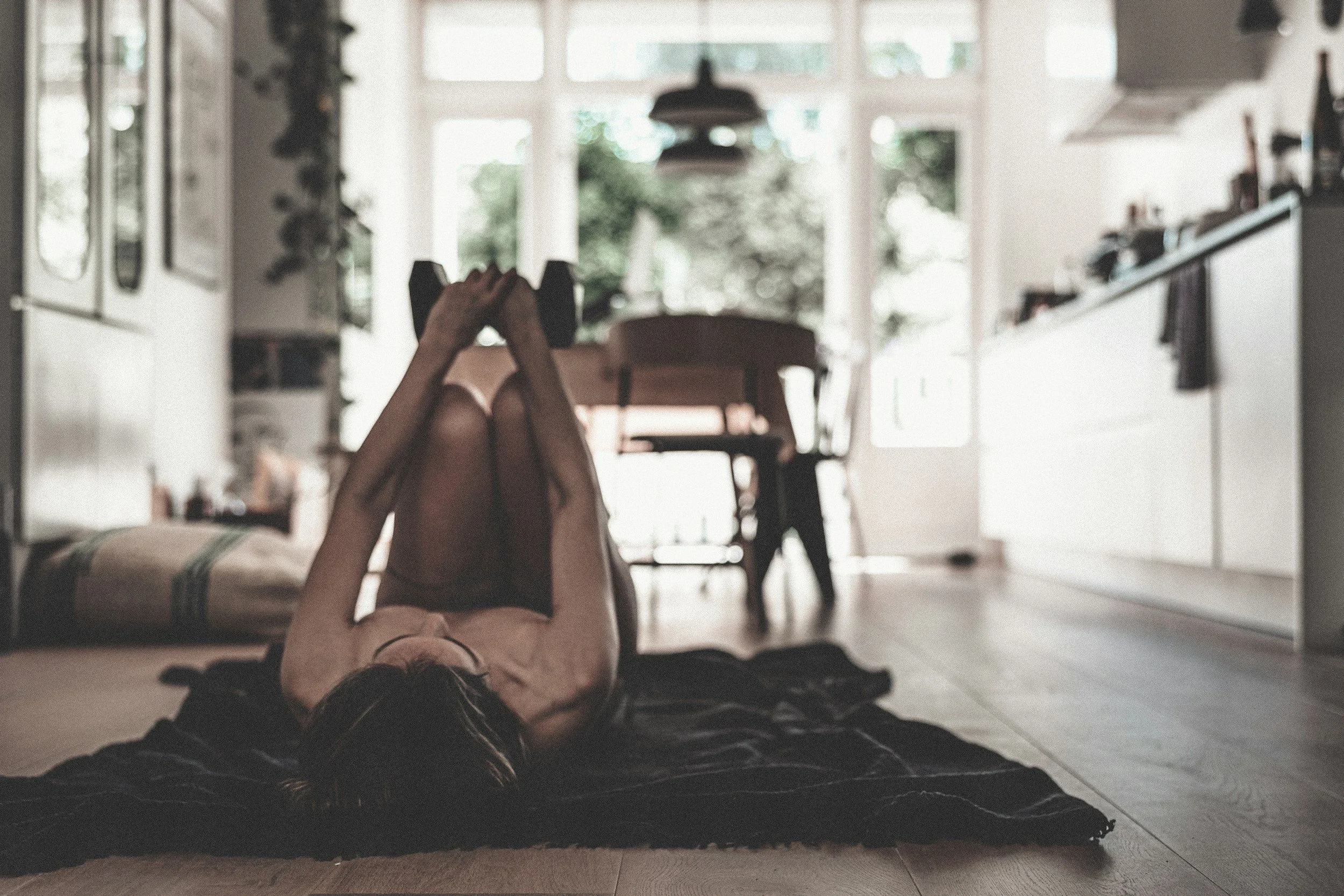 A person lying on their back on a rug with legs raised, holding dumbbells, in a bright room with large windows. The room has modern furniture and shelves, with a blurred view of a dining area and green trees outside.