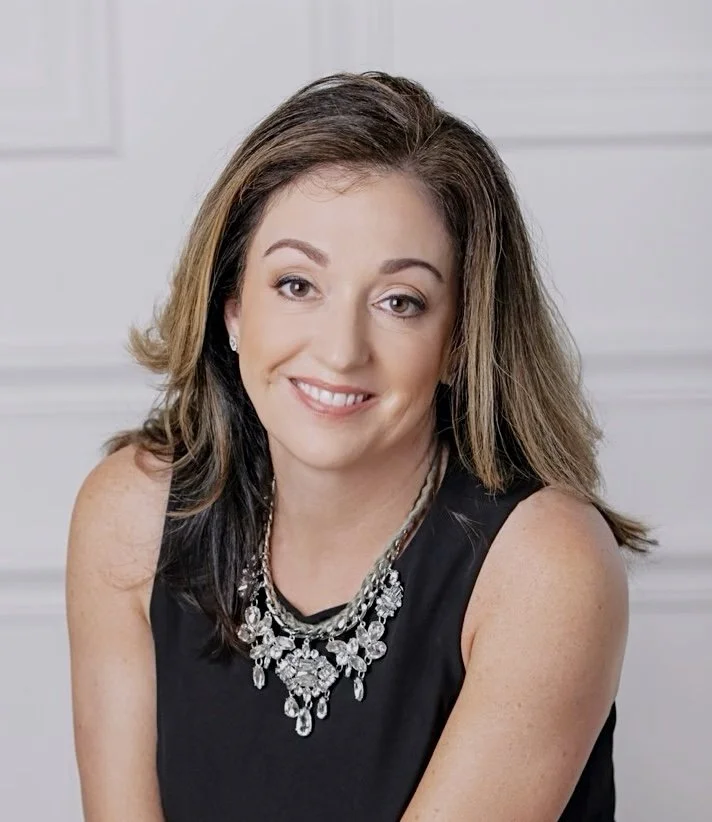 A smiling woman with shoulder-length brown hair and light skin, wearing a sleeveless black top and a large silver necklace, sitting in front of a white paneled wall.