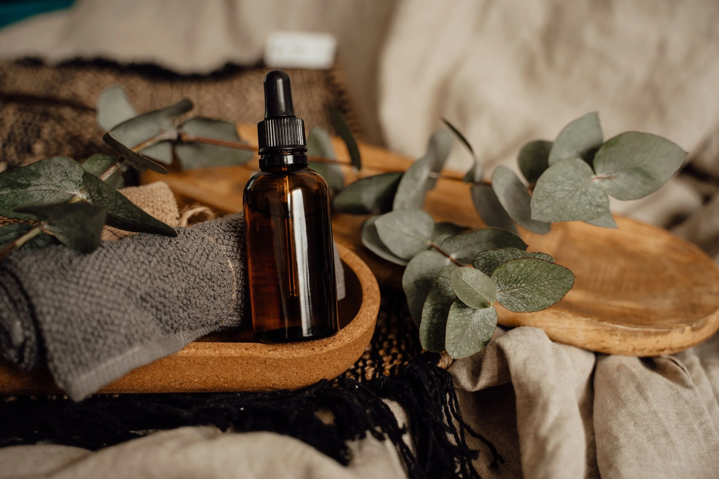 Amber glass dropper bottle placed among eucalyptus leaves, gray fabric, and textured materials on a wooden surface.
