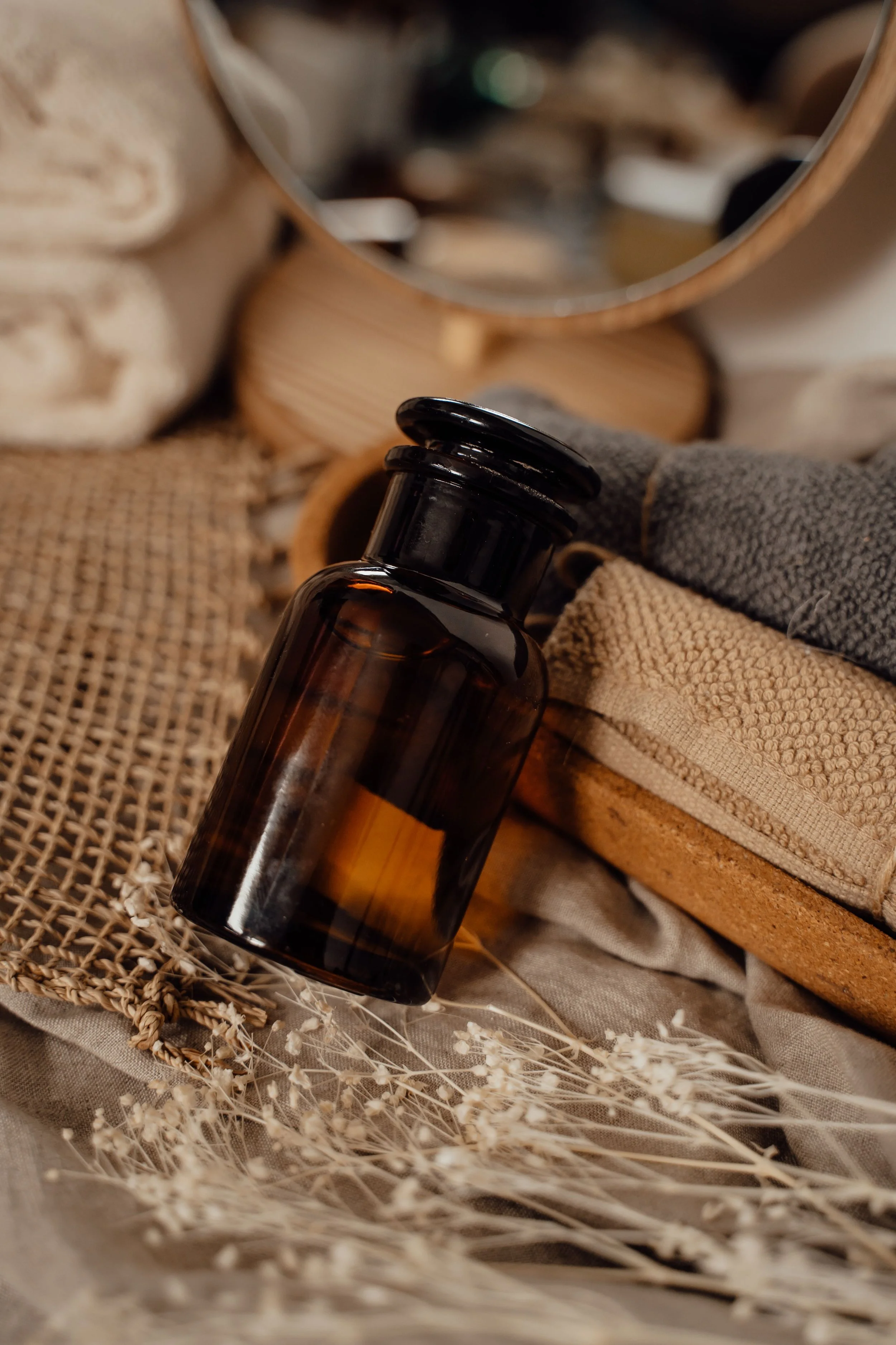 A dark amber glass bottle with a black cap placed among woven fabrics, dried plants, and a mirror reflecting a blurred background.