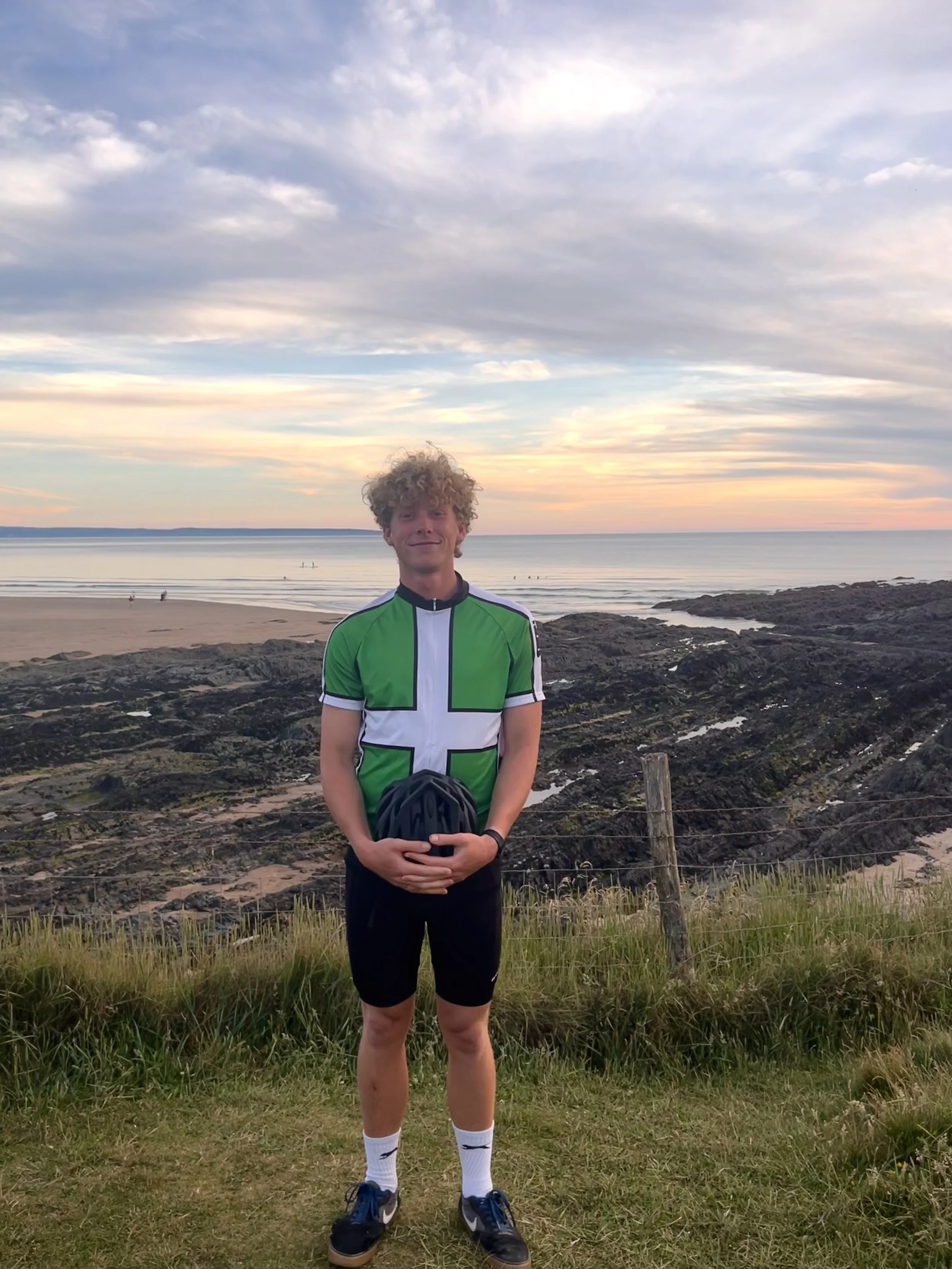 A young man with curly hair standing on grassy terrain by the ocean at sunset, holding a helmet and wearing a green and white sports jersey, black shorts, white socks, and running shoes.