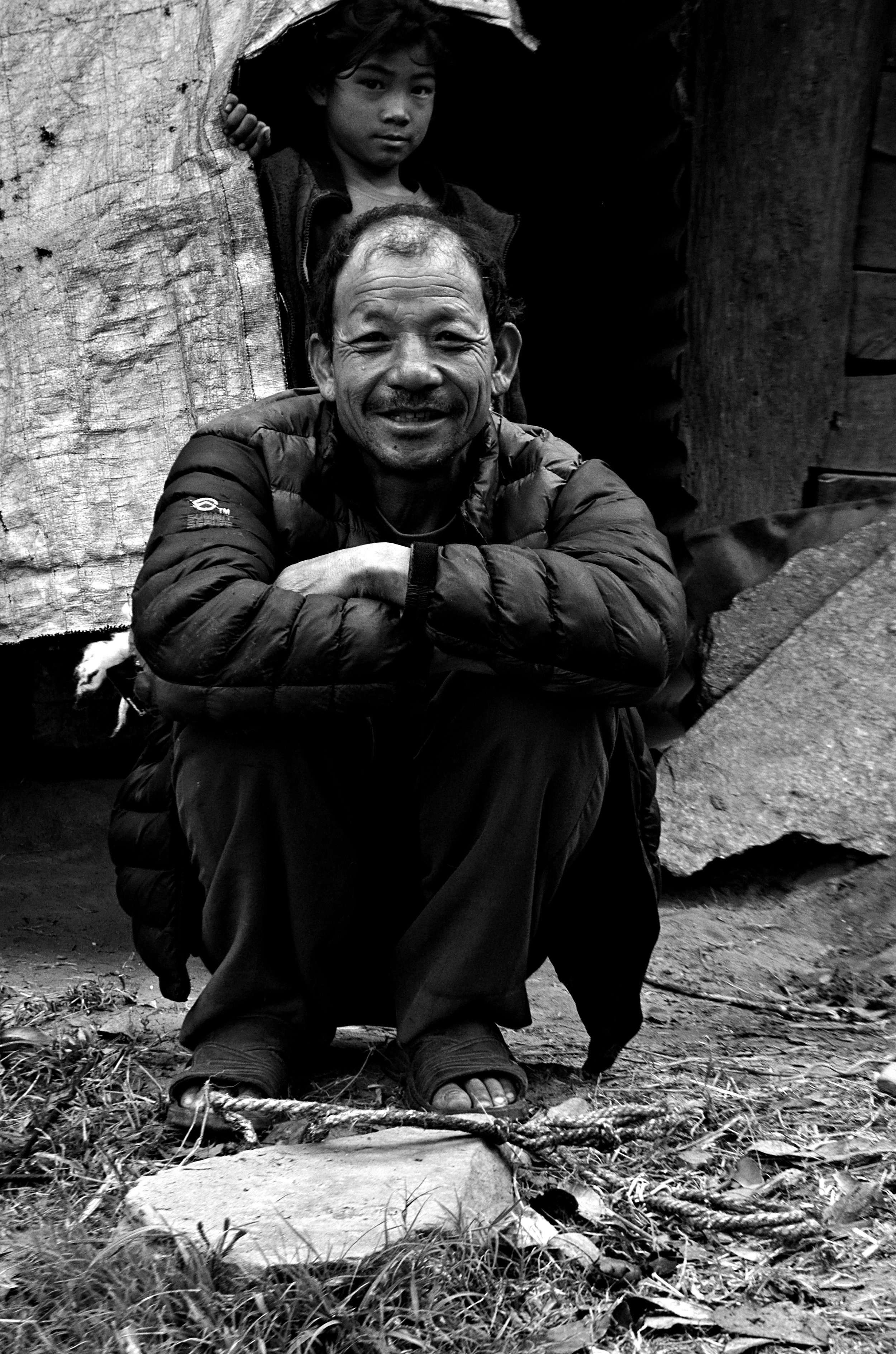 A black and white photo of a man and a boy. The man is sitting on the ground with his arms crossed on his knees, smiling at the camera, wearing a puffy jacket and sandals. The boy is standing behind him, peering out from a dark hole in a wooden structure, with a serious expression.