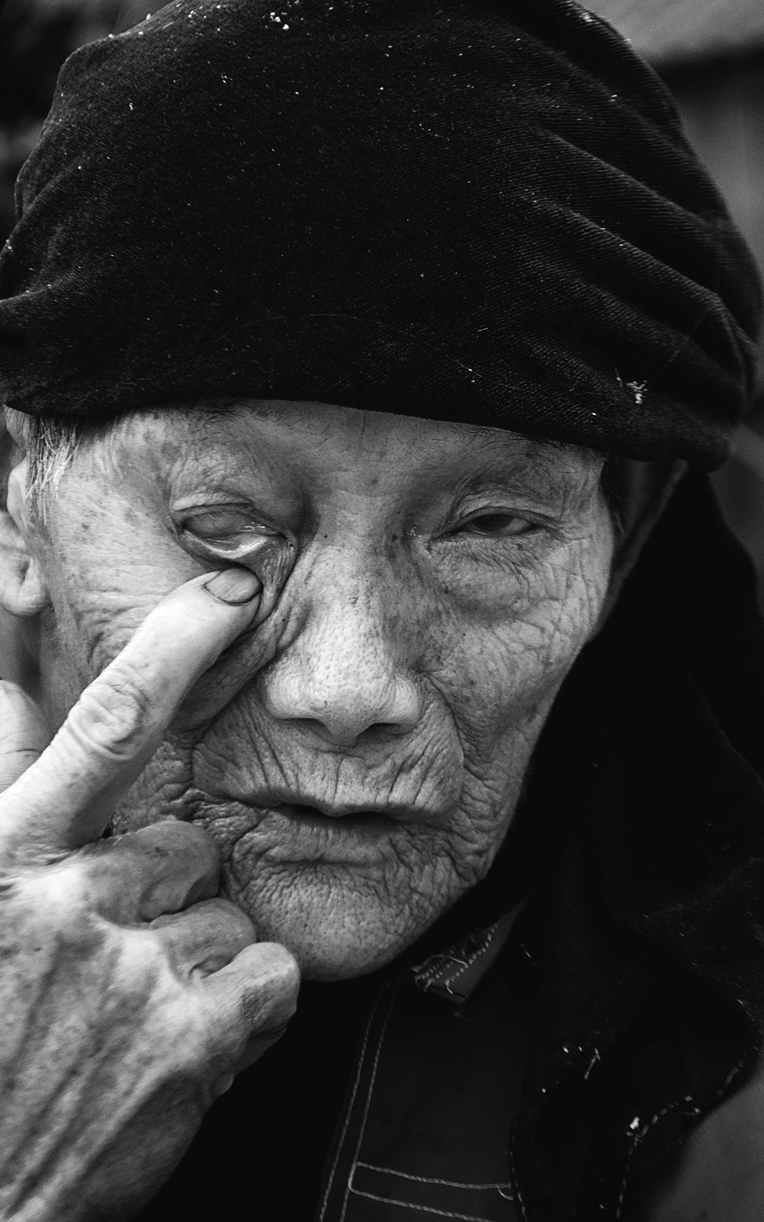 A close-up black-and-white photo of an elderly person wearing a dark hat. The person is touching their face with one hand, with their index finger resting near their eye. The person's face shows signs of aging, with wrinkles and sagging skin.
