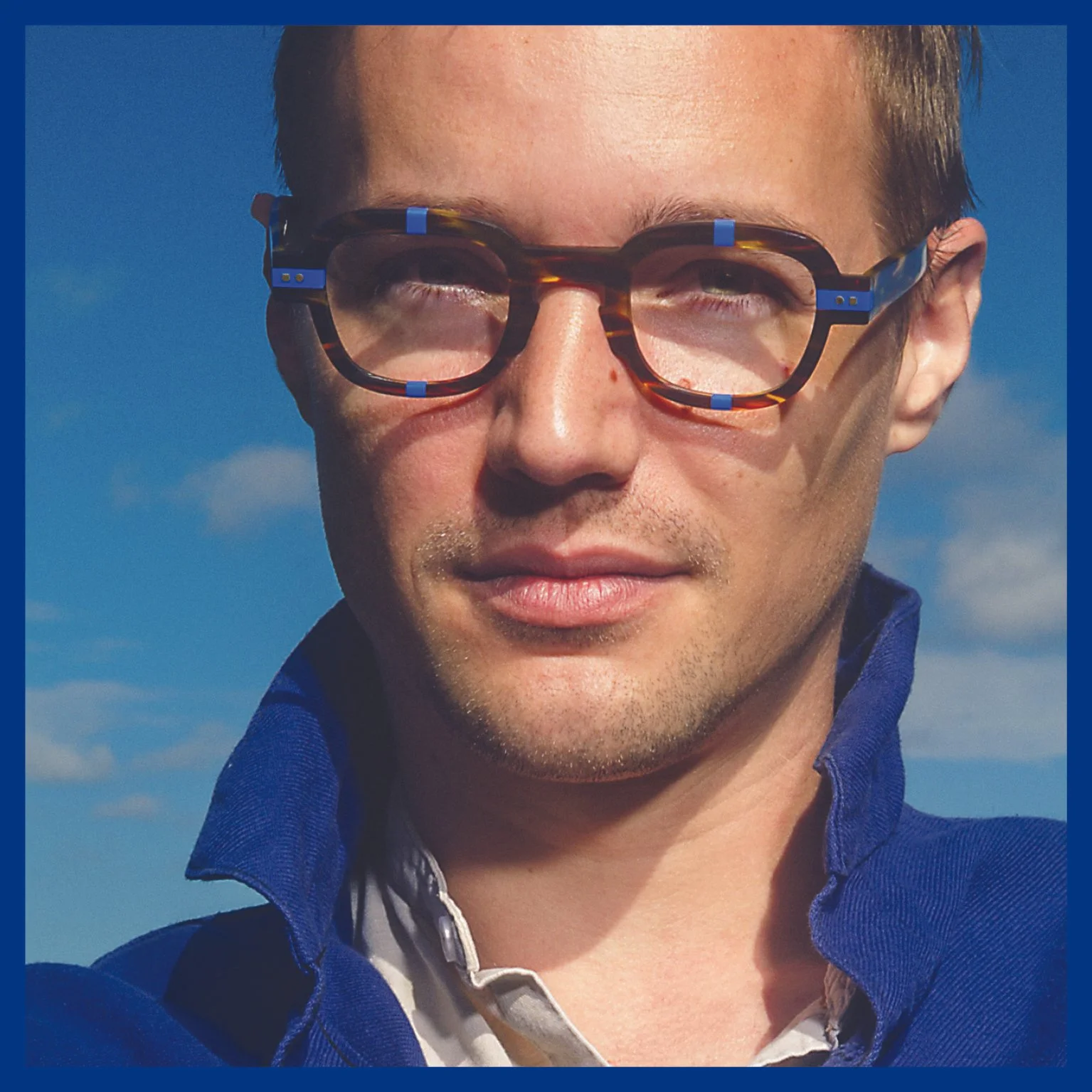 Close-up of a man with light skin and short brown hair wearing tortoiseshell glasses with blue accents, a blue jacket, and a white collared shirt underneath, standing outdoors against a blue sky with some clouds.