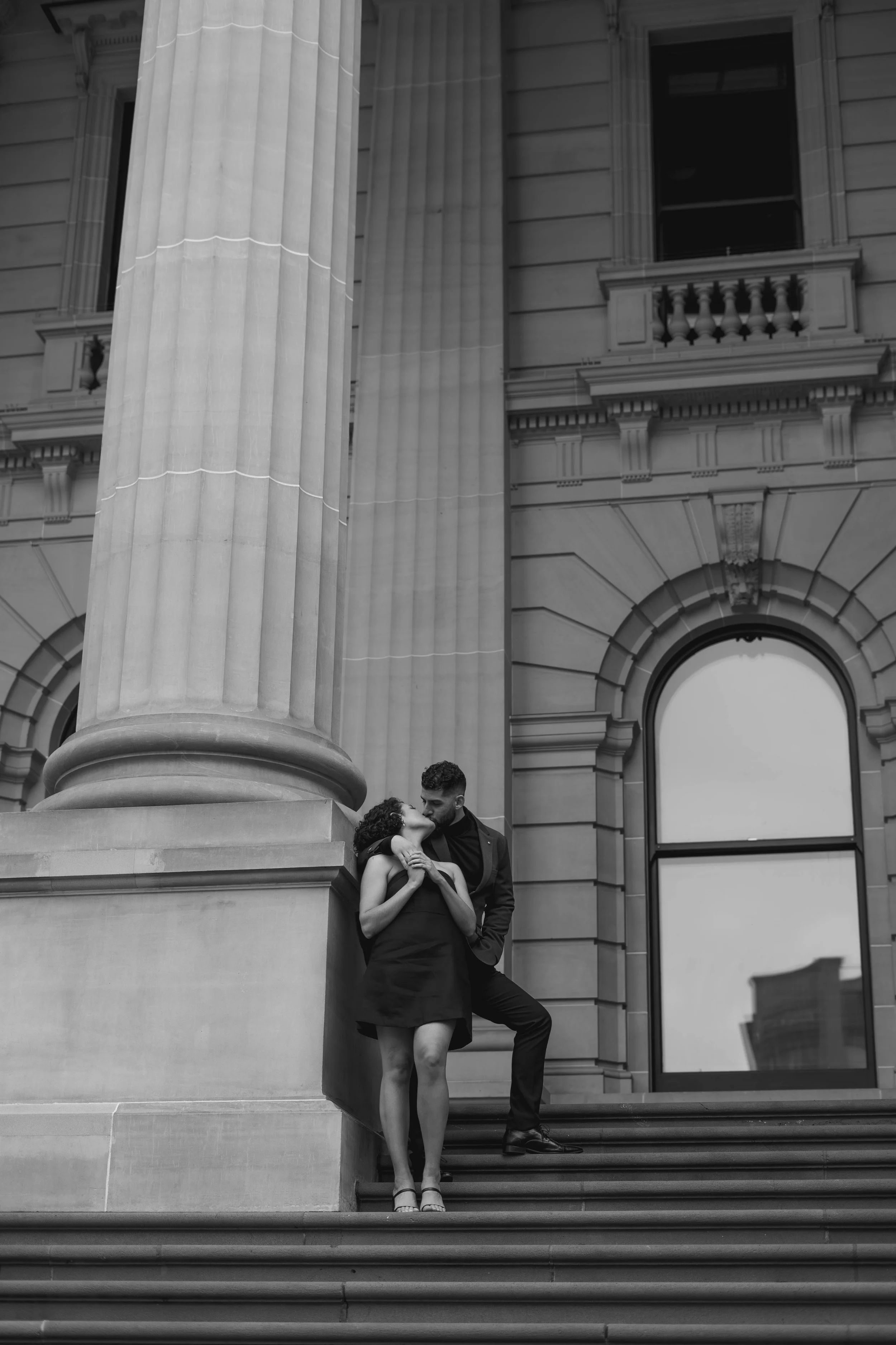 A black and white photo of a man and woman embracing on the steps of a large classical building with columns, arched windows, and decorative architectural details.