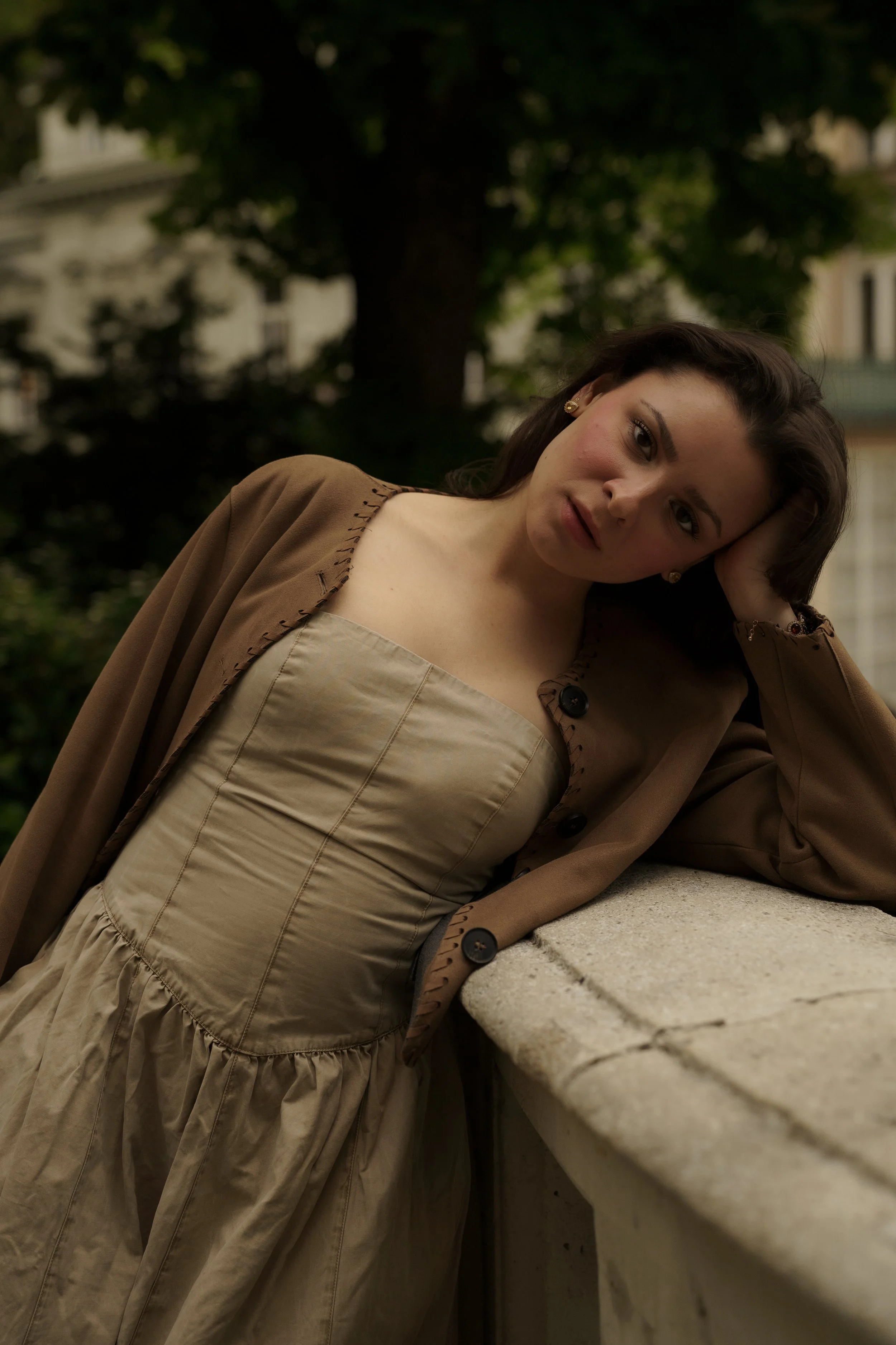 A woman leaning on a stone ledge with her head resting on her hand, dressed in a beige dress with a brown coat, outdoors with trees and buildings in the background.