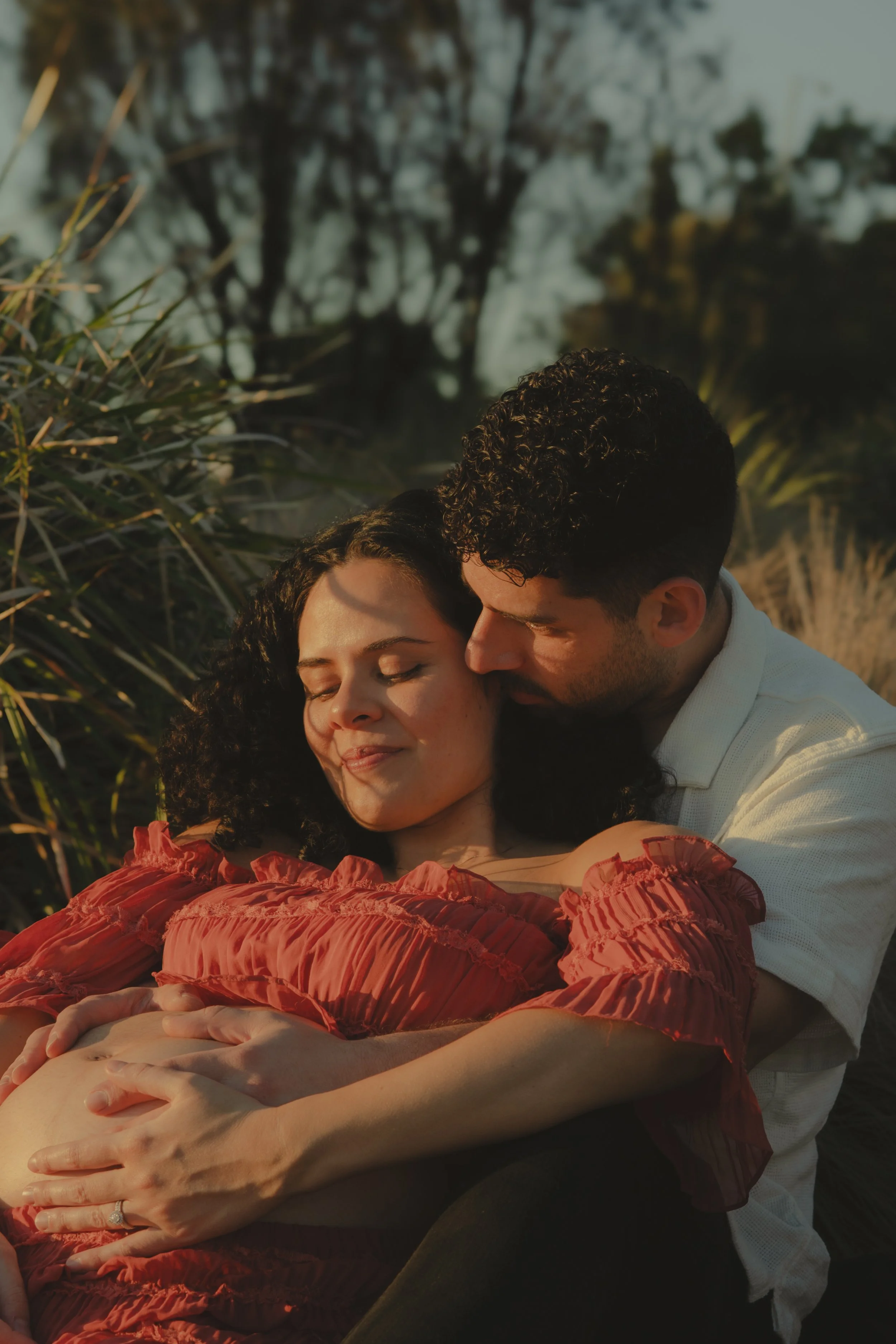 A couple embracing outdoors during sunset, with their eyes closed, surrounded by greenery and trees in the background.