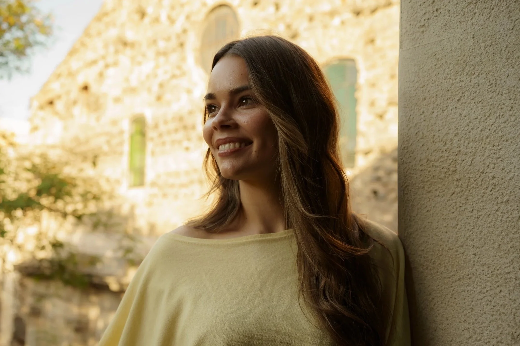 A woman with long brown hair and a nose piercing smiling while leaning against a beige wall outdoors with an old stone building in the background, during daylight.