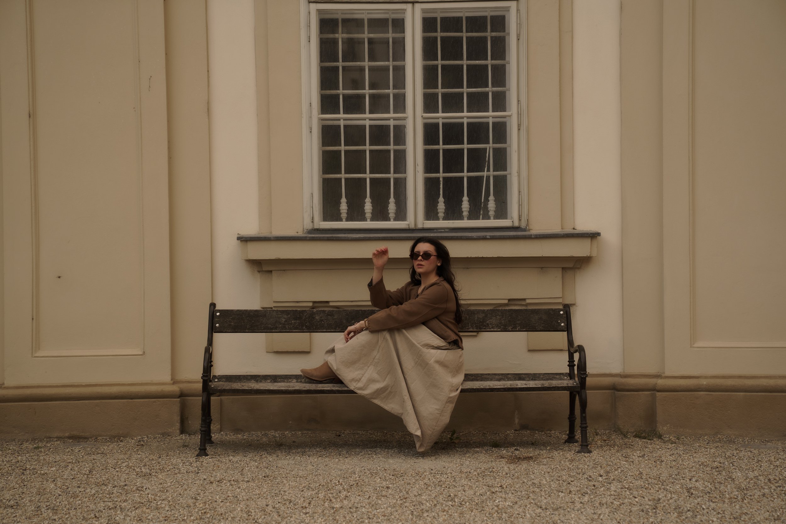 Woman sitting on a park bench in front of a large window and cream-colored wall, wearing sunglasses, a brown jacket, beige skirt, and boots, raising her right hand.