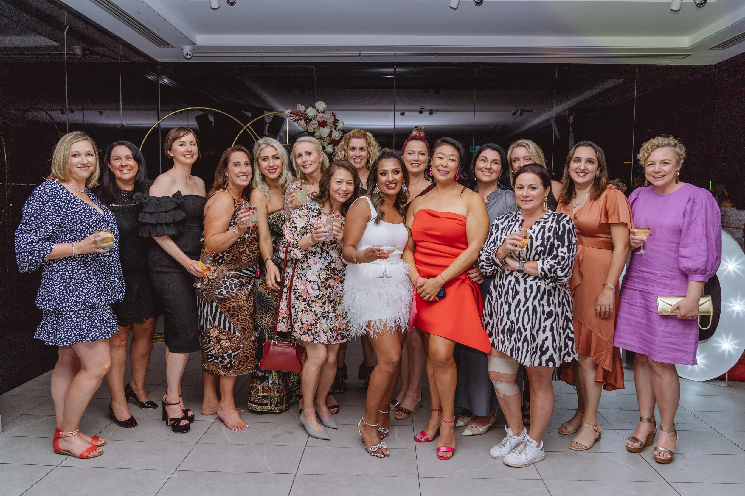 Group of women dressed in colorful and stylish outfits, holding drinks, standing together in a celebratory setting with decorative backdrop.