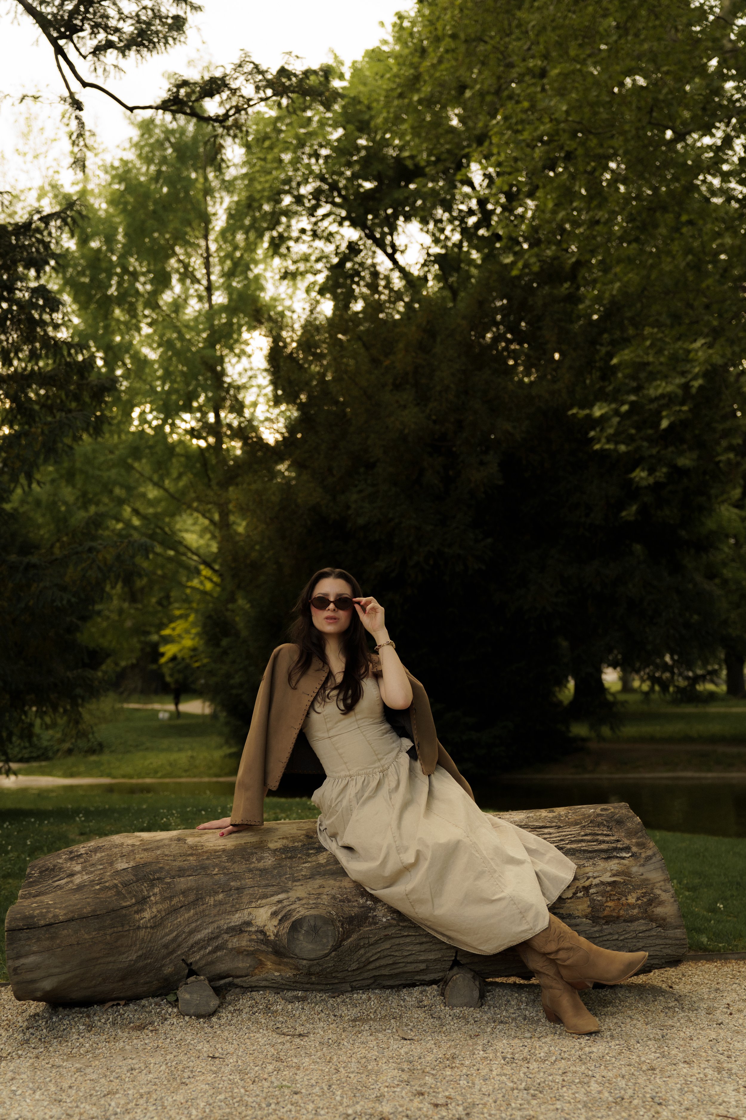 A woman wearing sunglasses, a beige dress, and boots sitting on a large fallen tree trunk in a park with trees and greenery in the background.