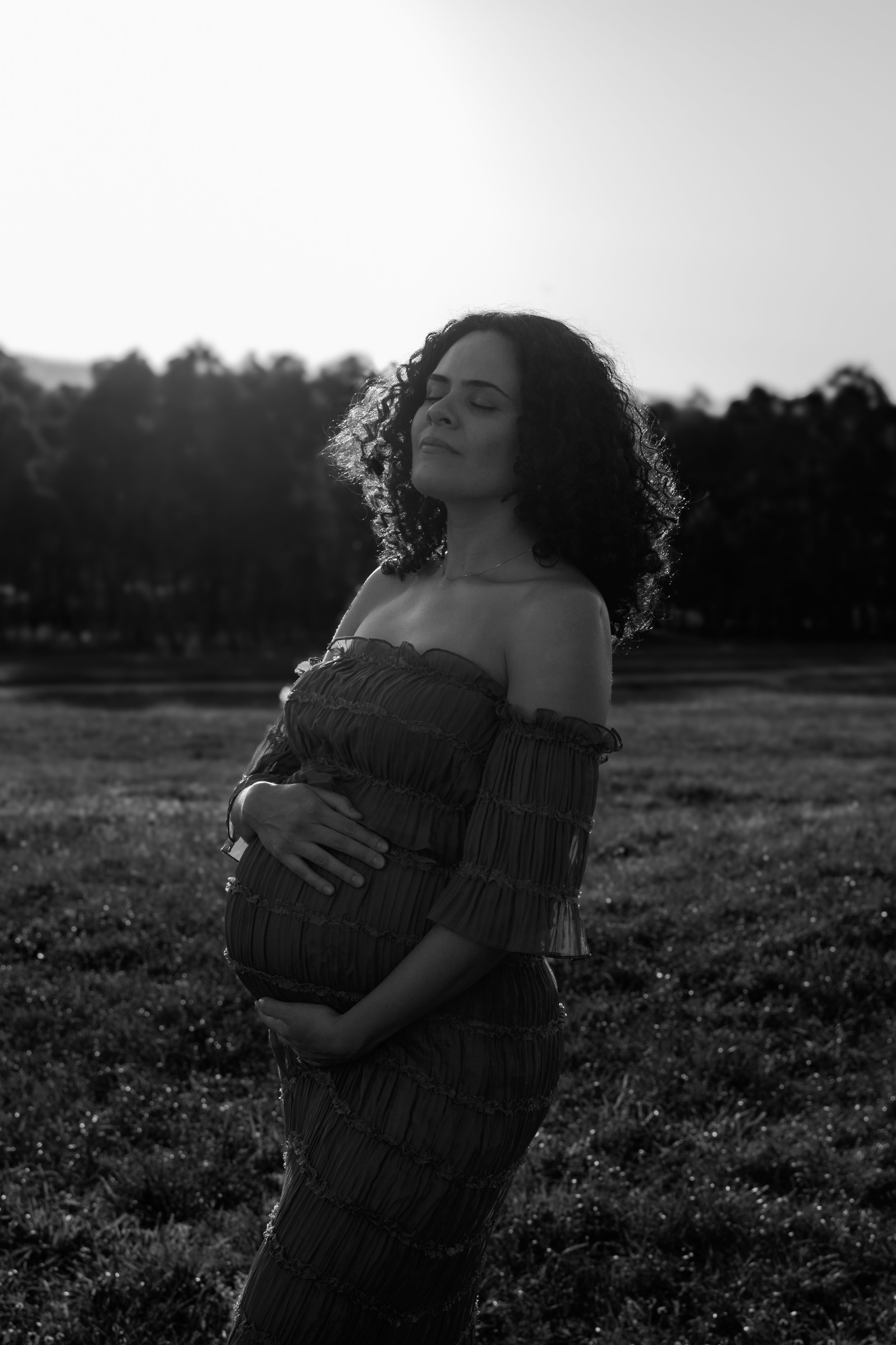 Black and white photo of a pregnant woman with curly hair standing outdoors with her eyes closed, gently holding her belly, with trees in the background.