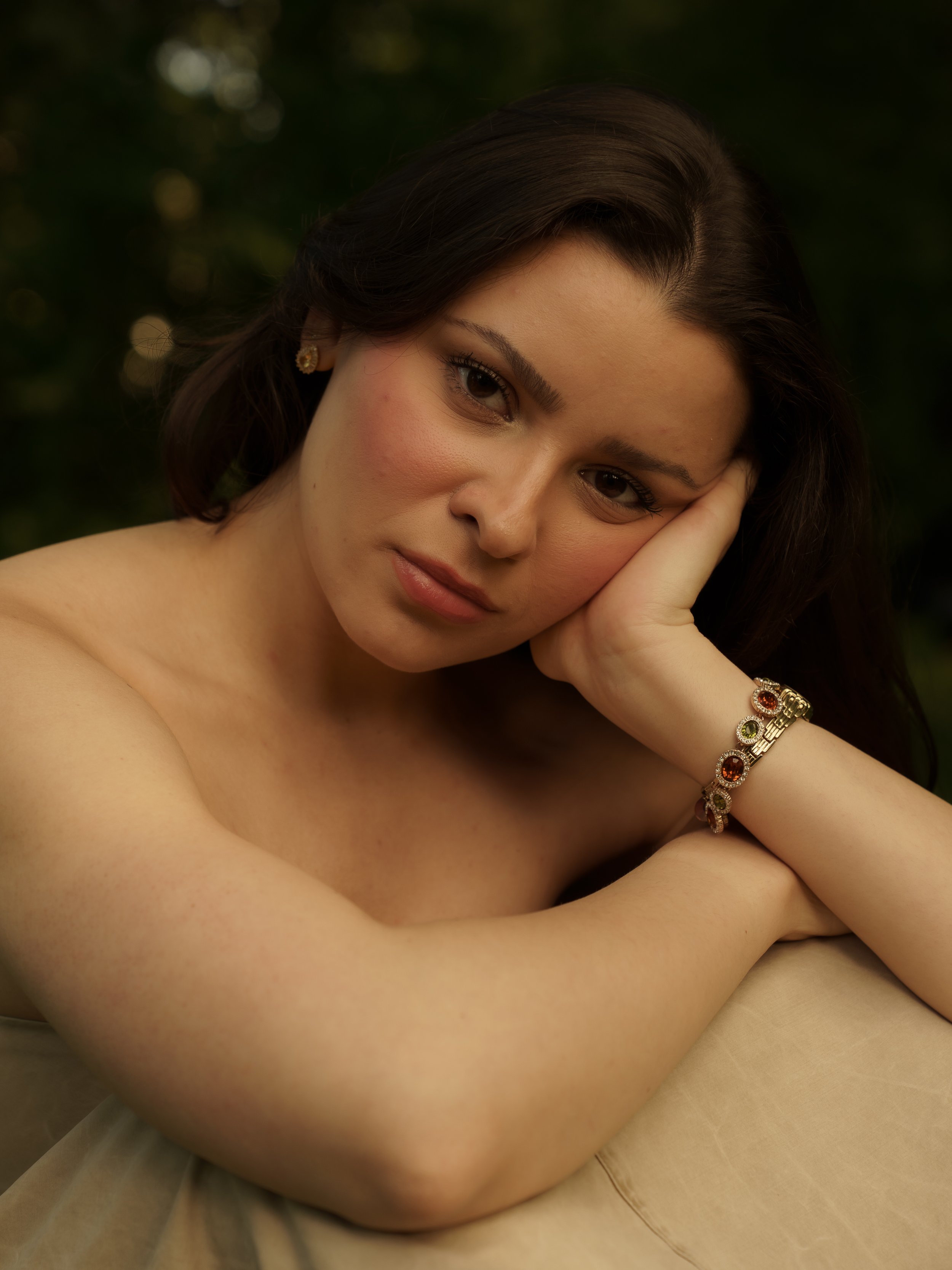 A woman with dark hair resting her head on her hand, wearing earrings and a bracelet, with a soft expression on her face against a blurred dark background.