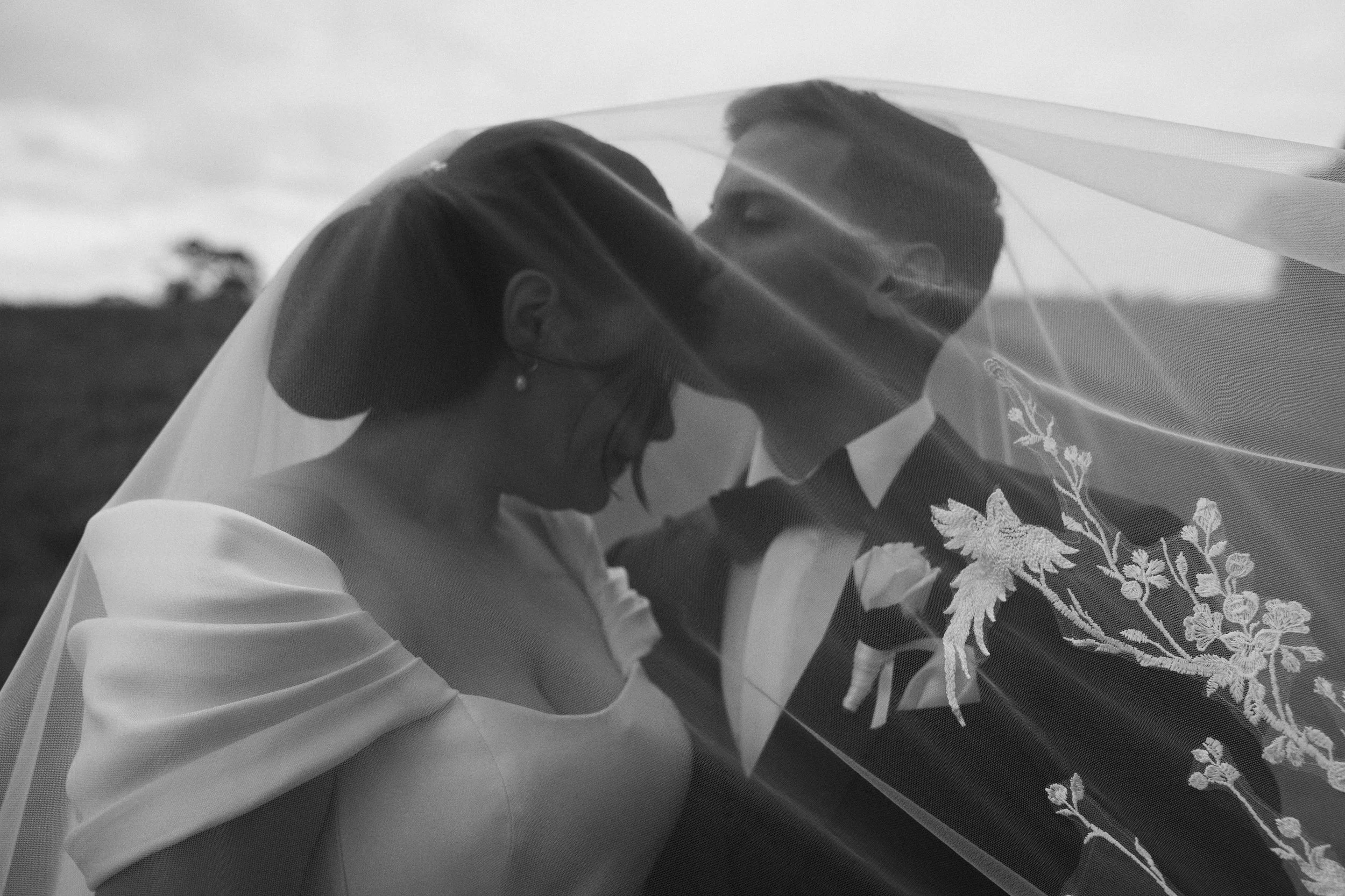 A man and woman on their wedding day, close together, sharing a kiss under a veil, outdoors with sky and trees in the background.