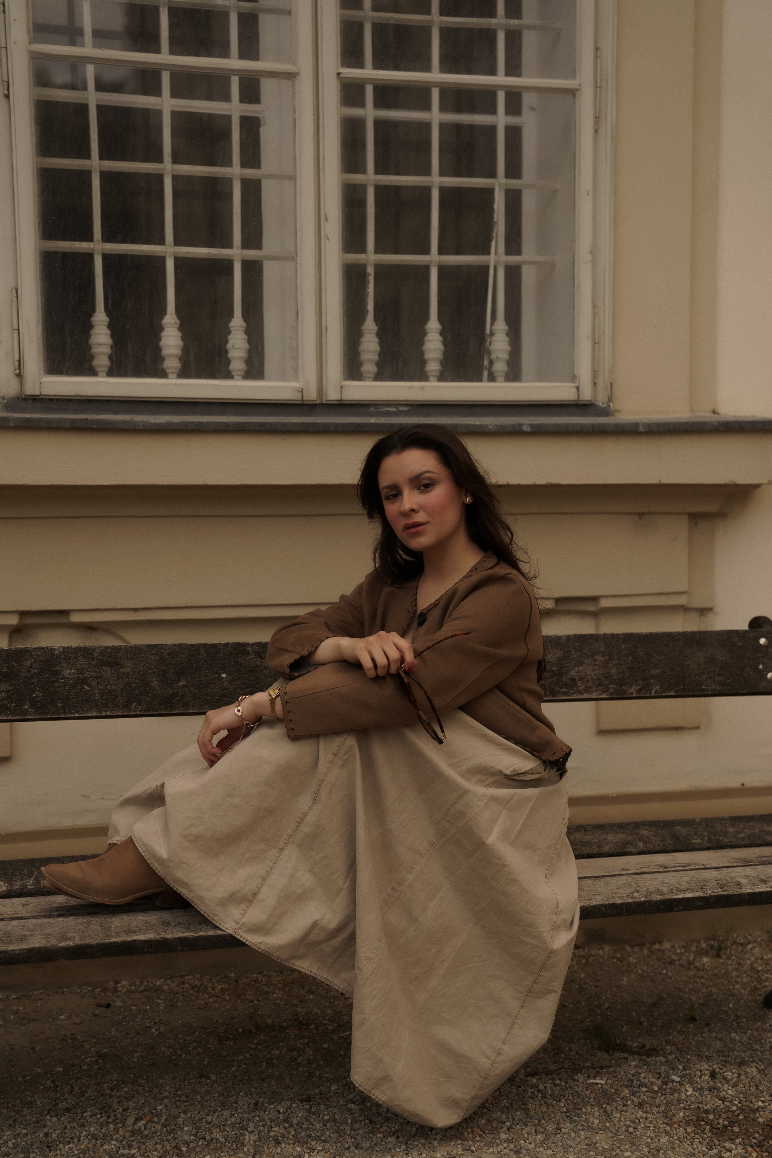 A woman with dark hair sitting on a weathered outdoor wooden bench with her legs crossed. She is wearing a brown jacket, beige skirt, and tan boots. She is looking at the camera with a relaxed expression. Behind her is a light-colored building wall a